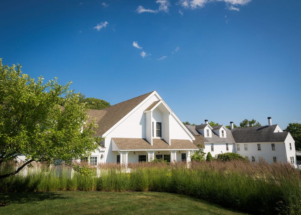 White farmhouse-style home surrounded by lush green landscaping under a bright blue summer sky.