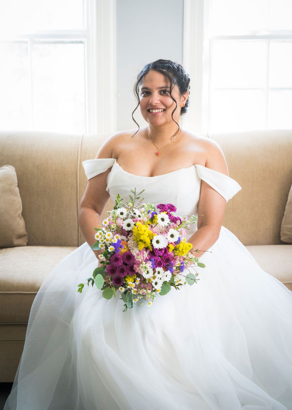 Smiling bride in off-shoulder white gown holds colorful wildflower bouquet while seated on a beige sofa.