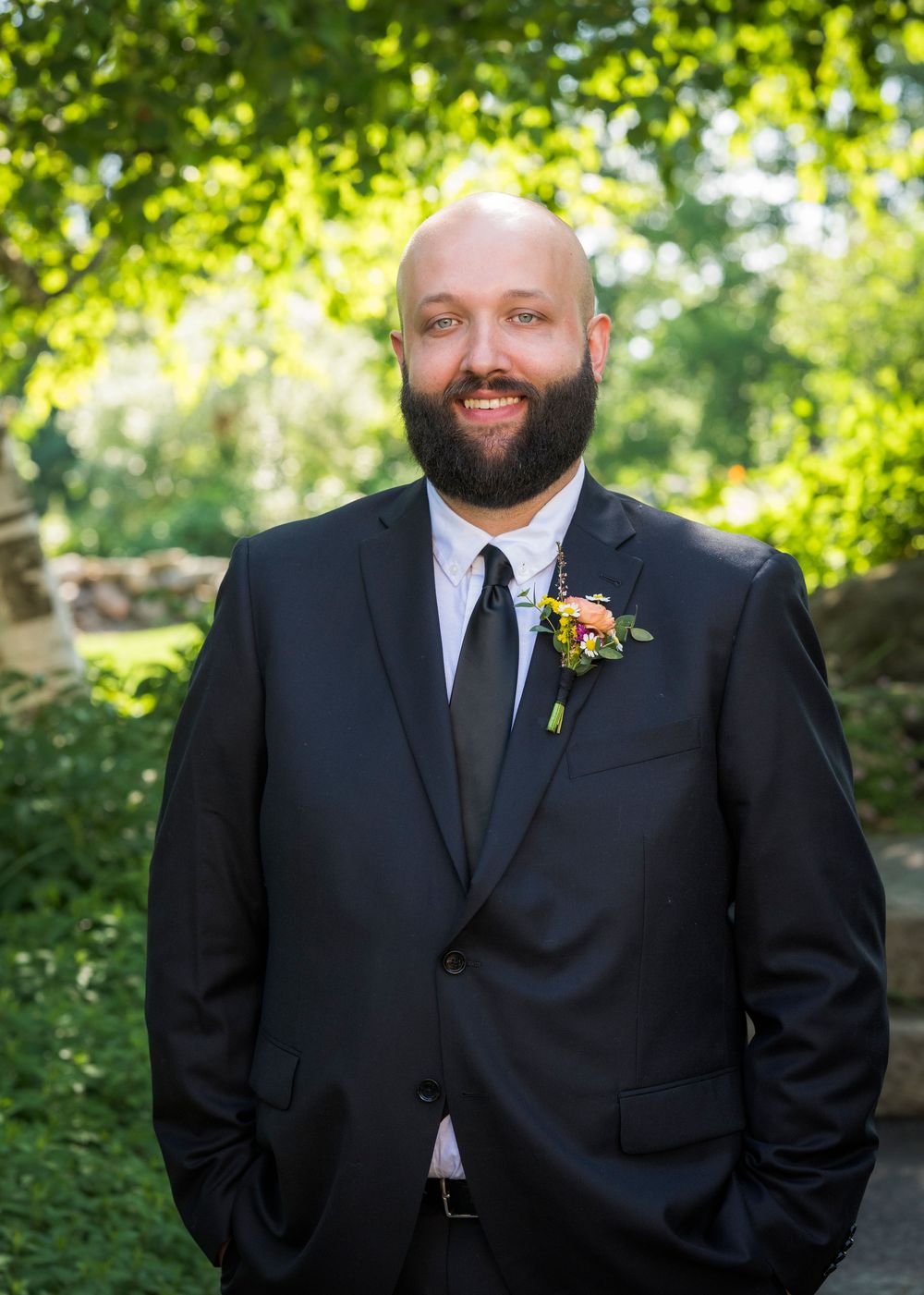 Smiling bald groom with beard wearing black suit and floral boutonniere standing outdoors.