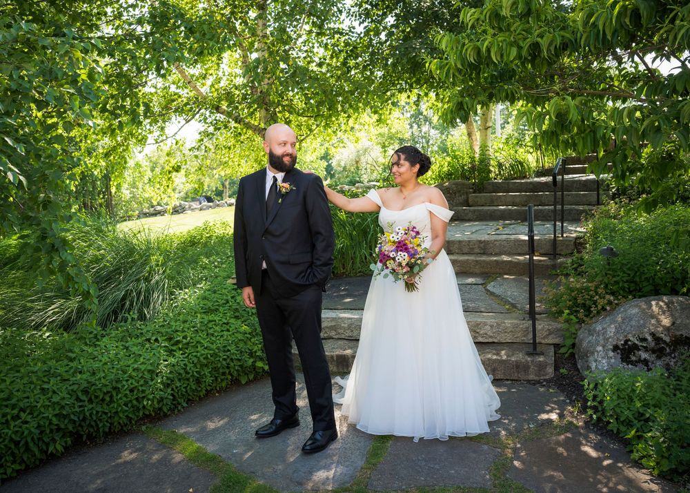 Bride in white gown touches groom's shoulder during outdoor wedding photo session in lush green garden.