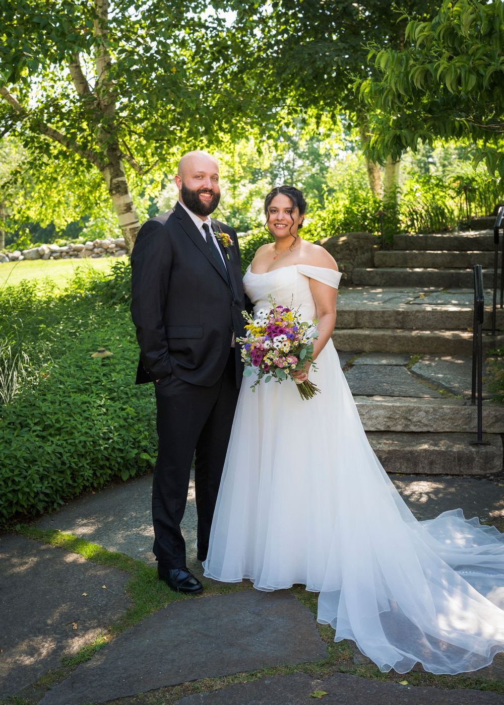 Bride and groom pose together in a lush garden setting, bride holding a colorful wildflower bouquet in a white gown.