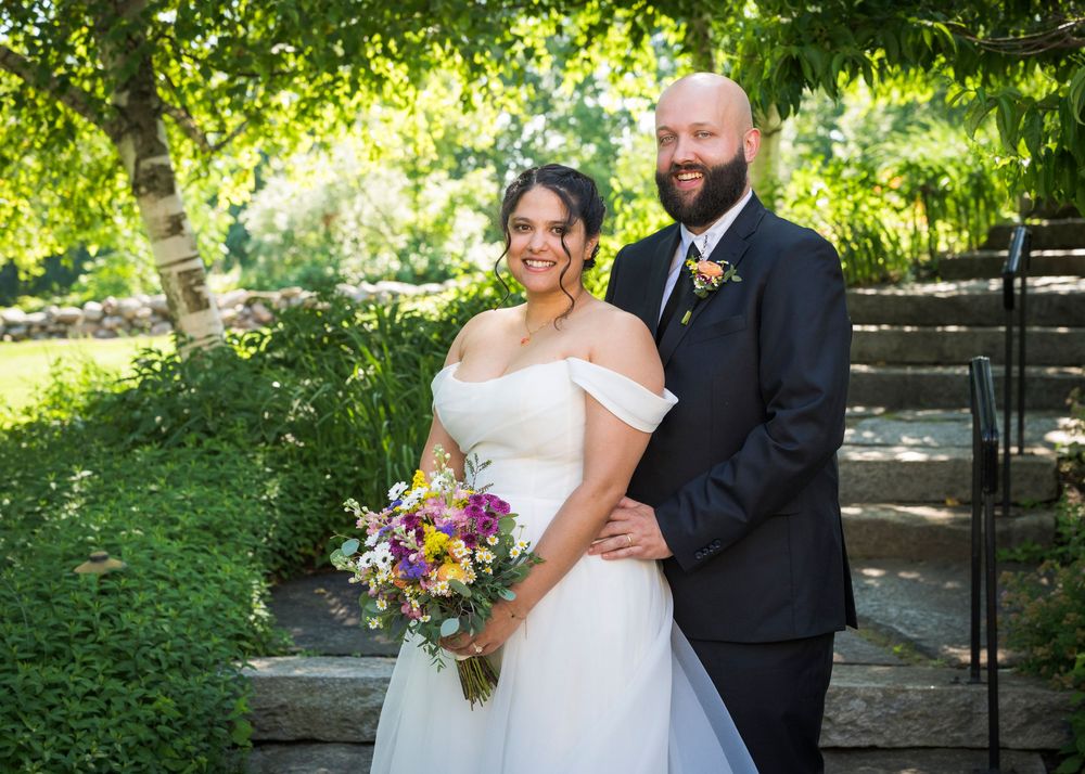 Bride in off-shoulder white gown holding colorful bouquet poses with bearded groom in black suit outdoors.