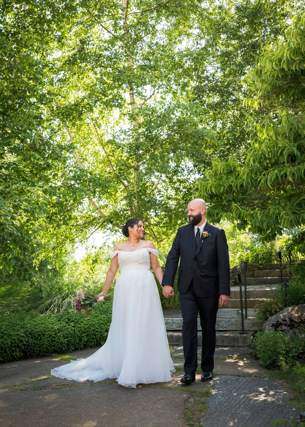 Bride and groom holding hands walking through a lush green garden on their wedding day.