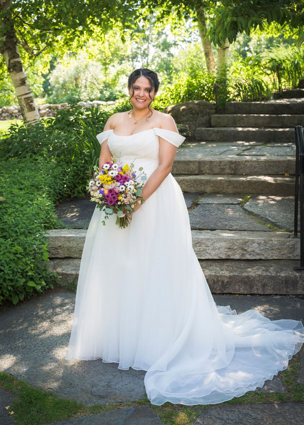 Bride in off-shoulder white ball gown with train holds colorful bouquet in a lush garden setting.