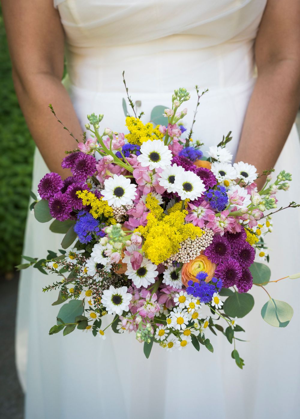 Bride holds a vibrant wildflower wedding bouquet featuring purple, yellow, white, and blue blooms with eucalyptus greenery.