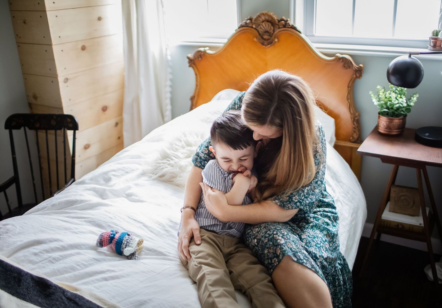 A laughing preschool boy is snuggled tightly by his blond mother wearing green floral dress in his bedroom with an antique bed in Groton MA.
