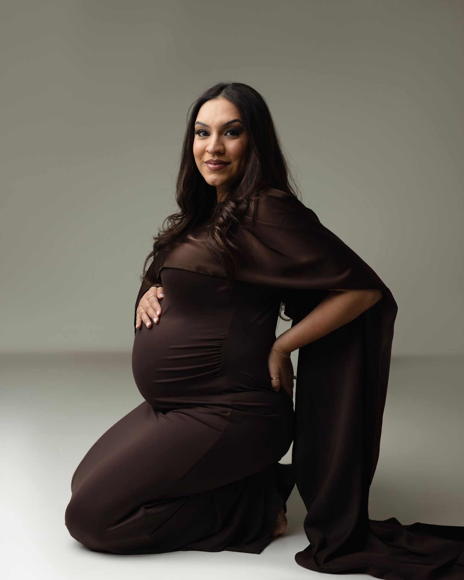 Woman in brown dress poses gracefully in maternity photoshoot against neutral backdrop. Elizabeth Lang Photography Surrey