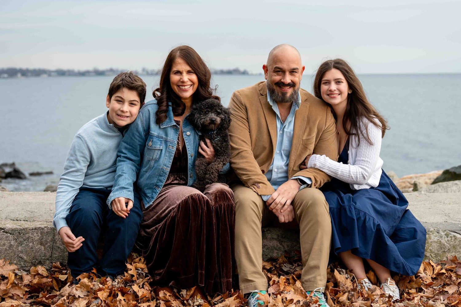 Family wearing coordinated blue and brown outfits poses together on autumn beach with fallen leaves.