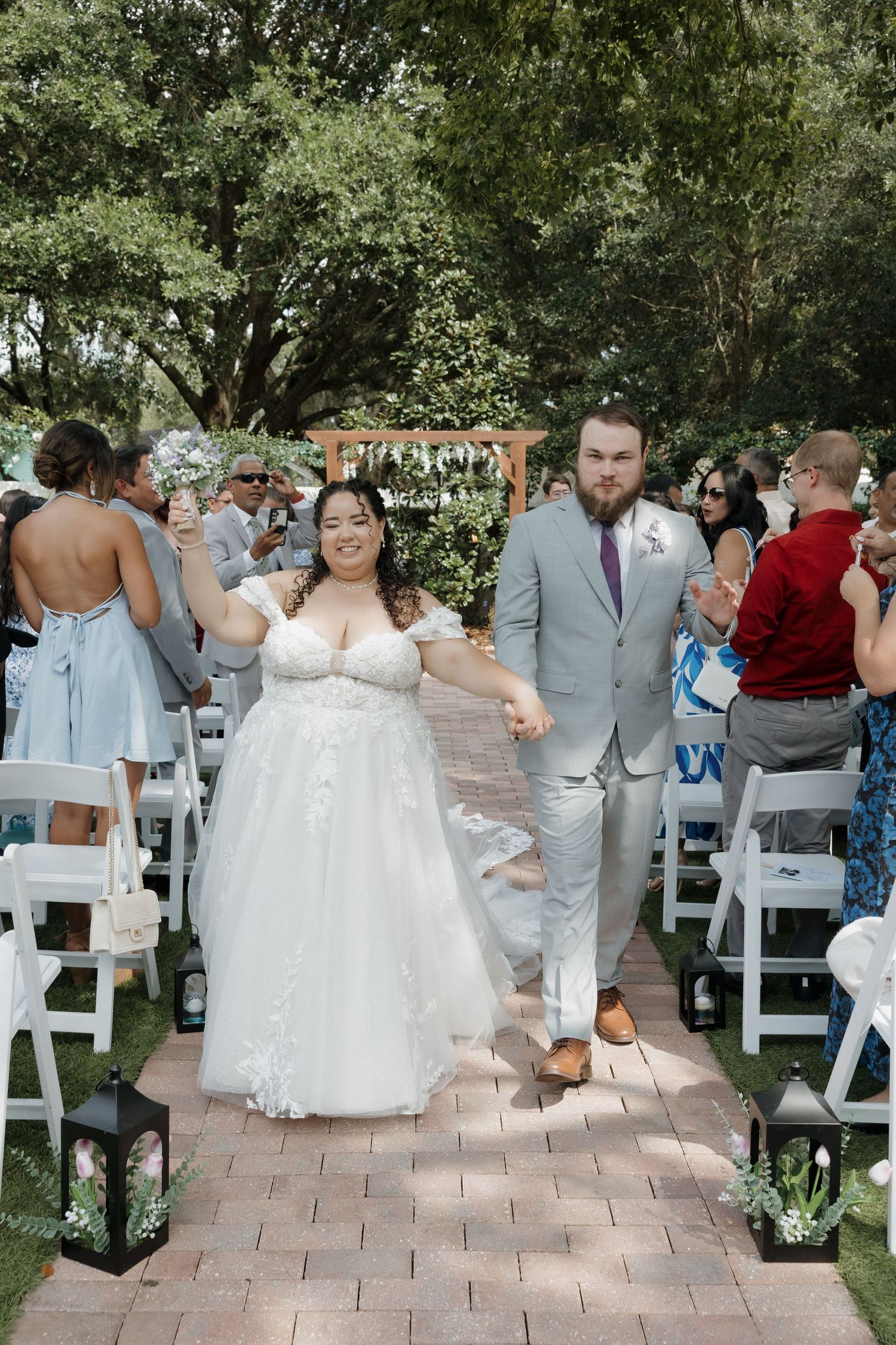 Newlyweds joyfully walk down outdoor aisle after ceremony surrounded by guests and lush green trees.