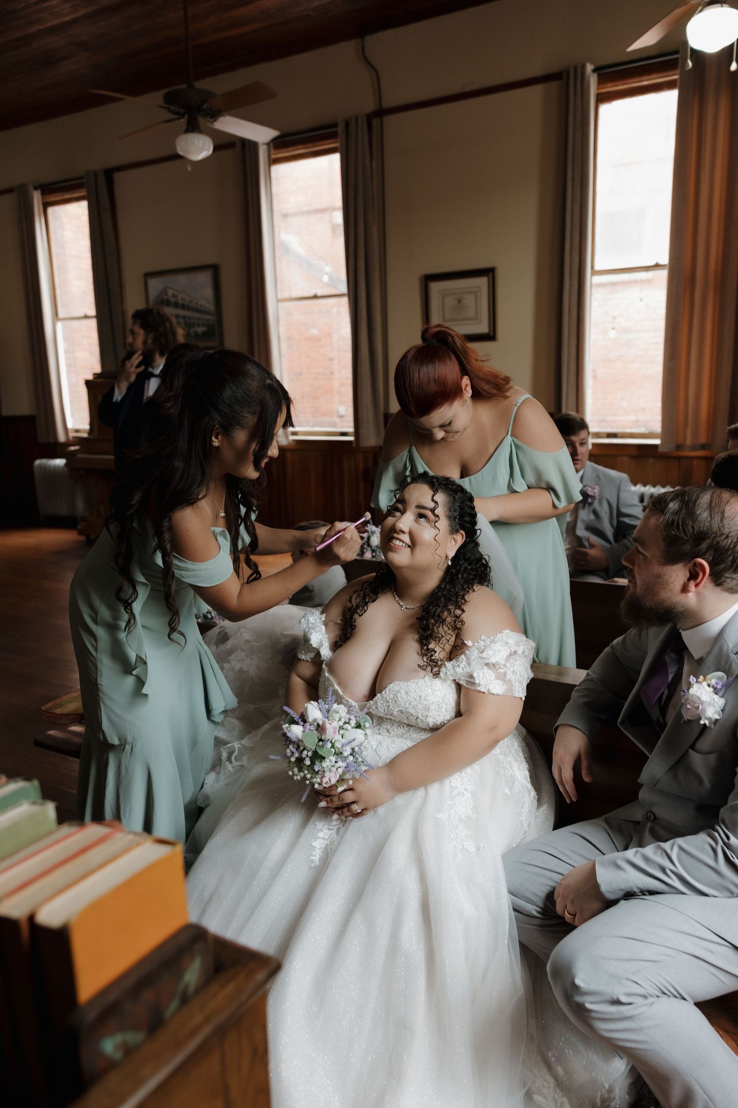 Bride in lace off-shoulder gown gets makeup done by bridesmaids in sage green dresses indoors.
