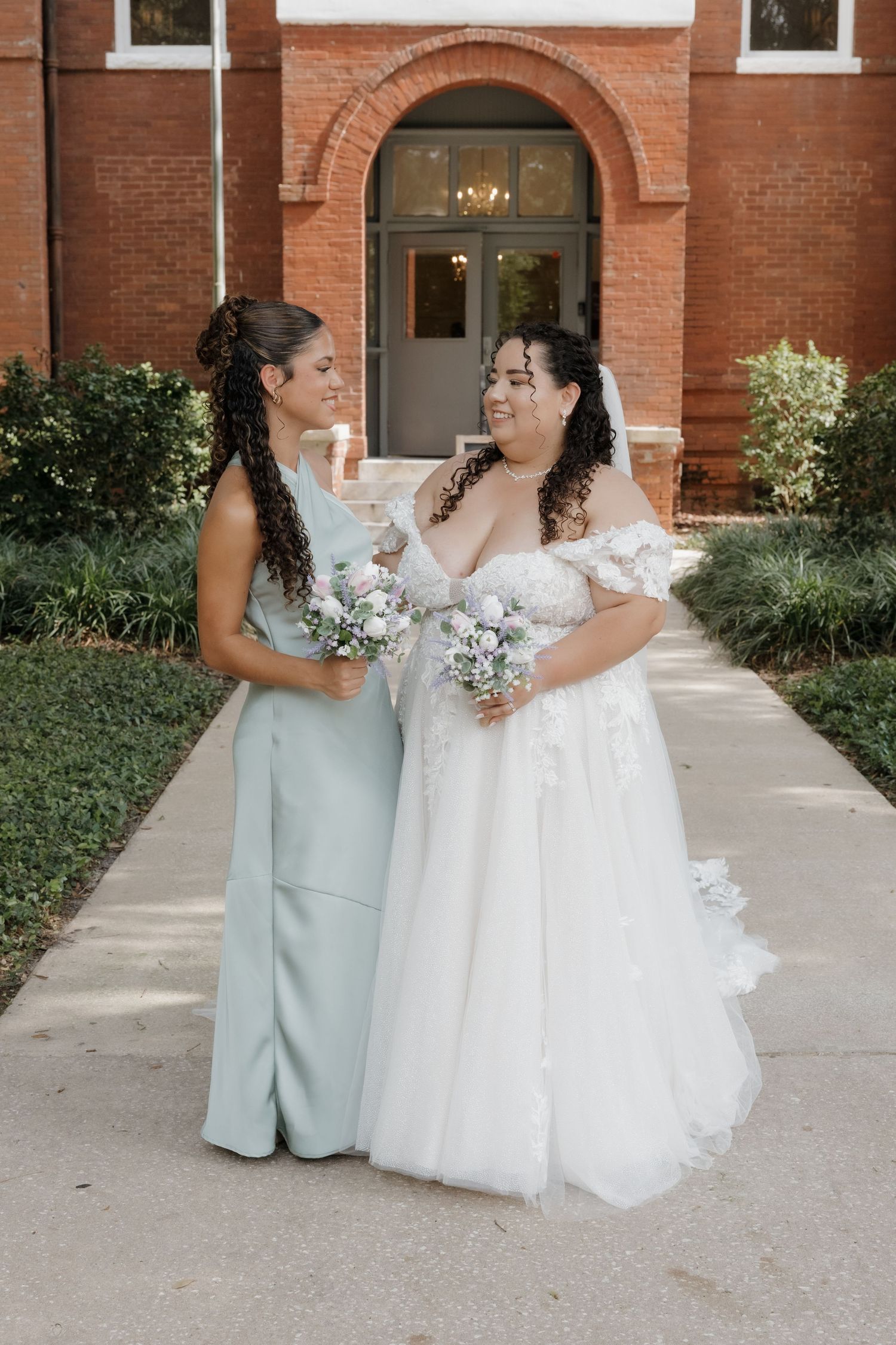 Bride in off-shoulder floral gown walks with bridesmaid in sage pantsuit outside brick building.