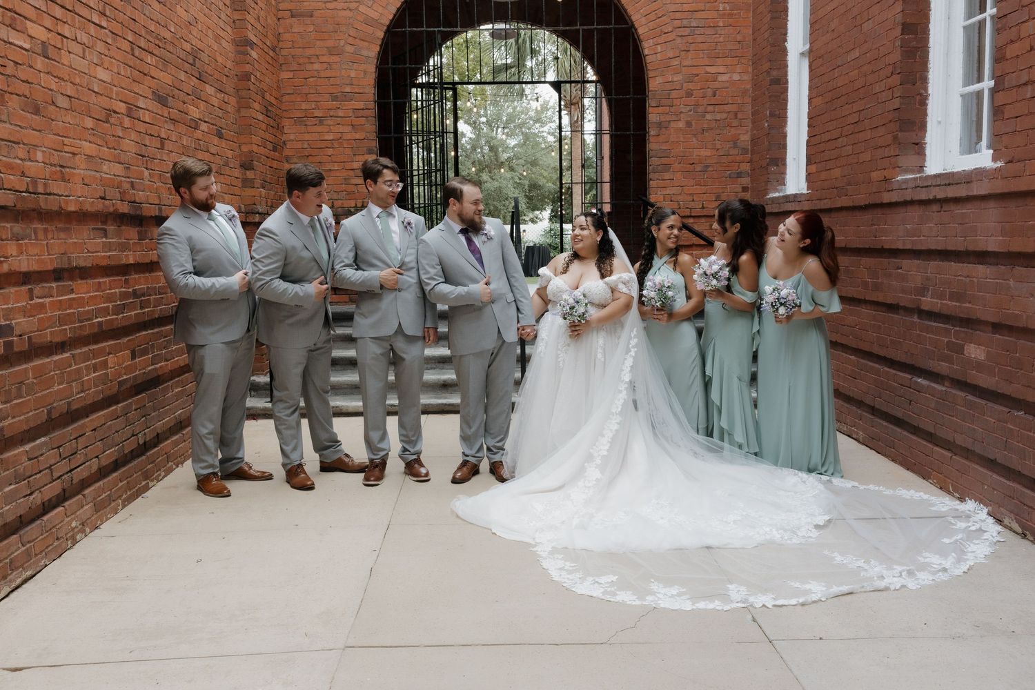 Wedding party posing in brick archway with bride in white gown and bridesmaids in sage green dresses.