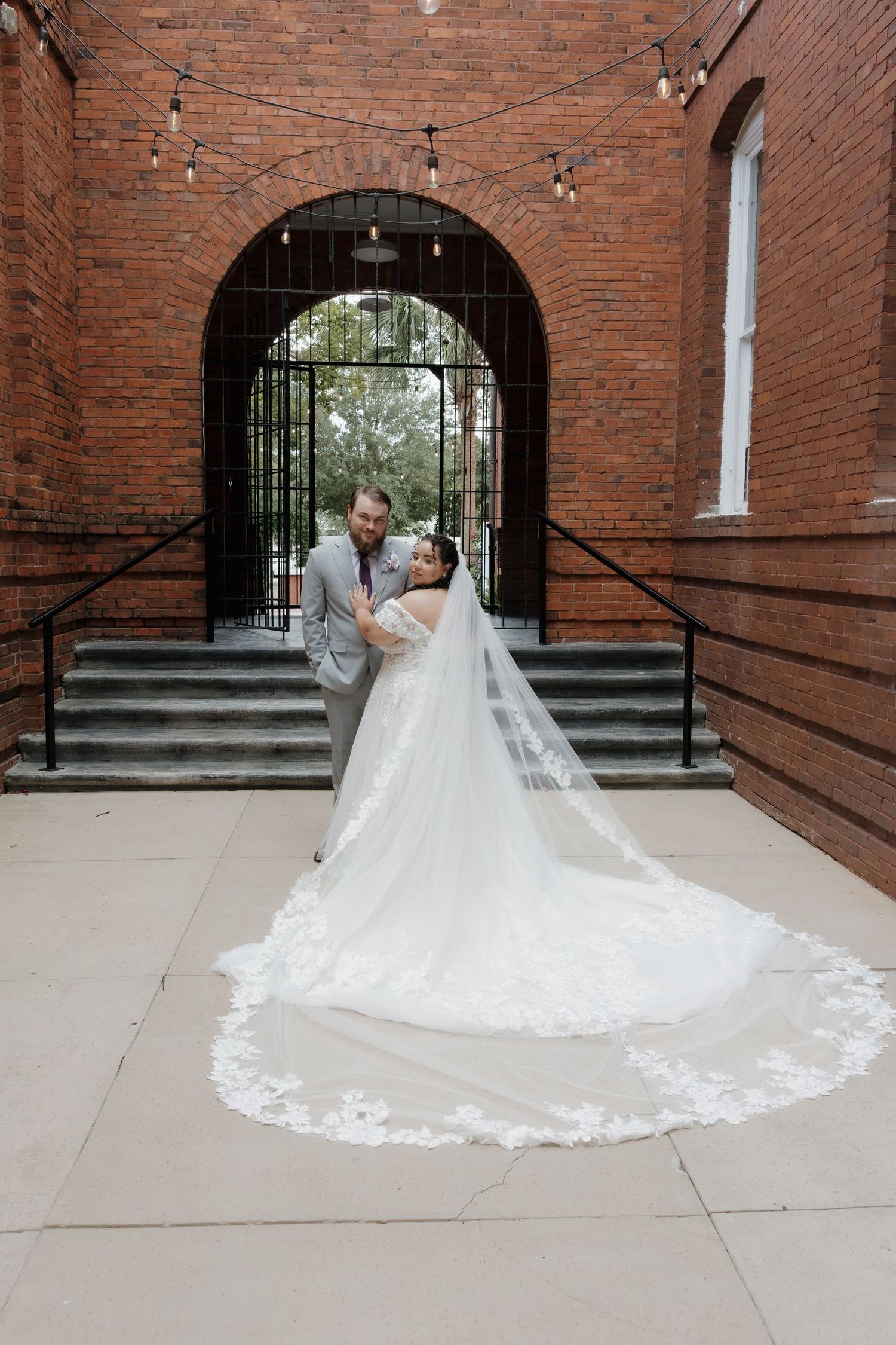 Bride and groom pose before brick arch with lace cathedral veil trailing on ground at elegant wedding venue.