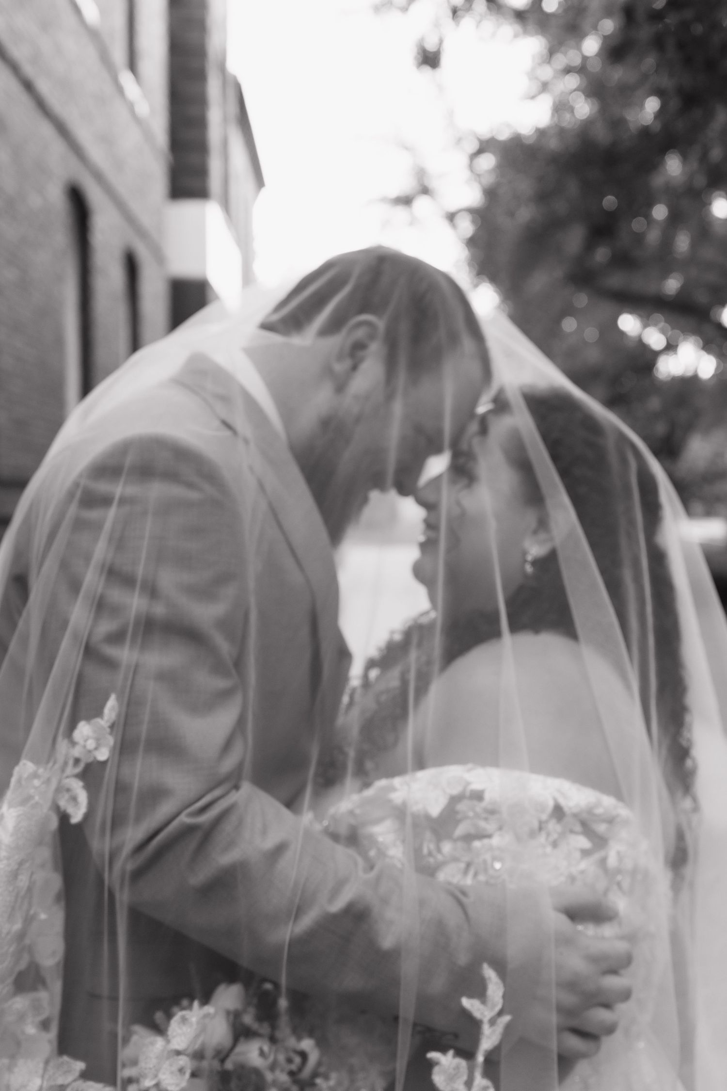 Black and white photo of bride and groom sharing intimate moment under flowing lace wedding veil.