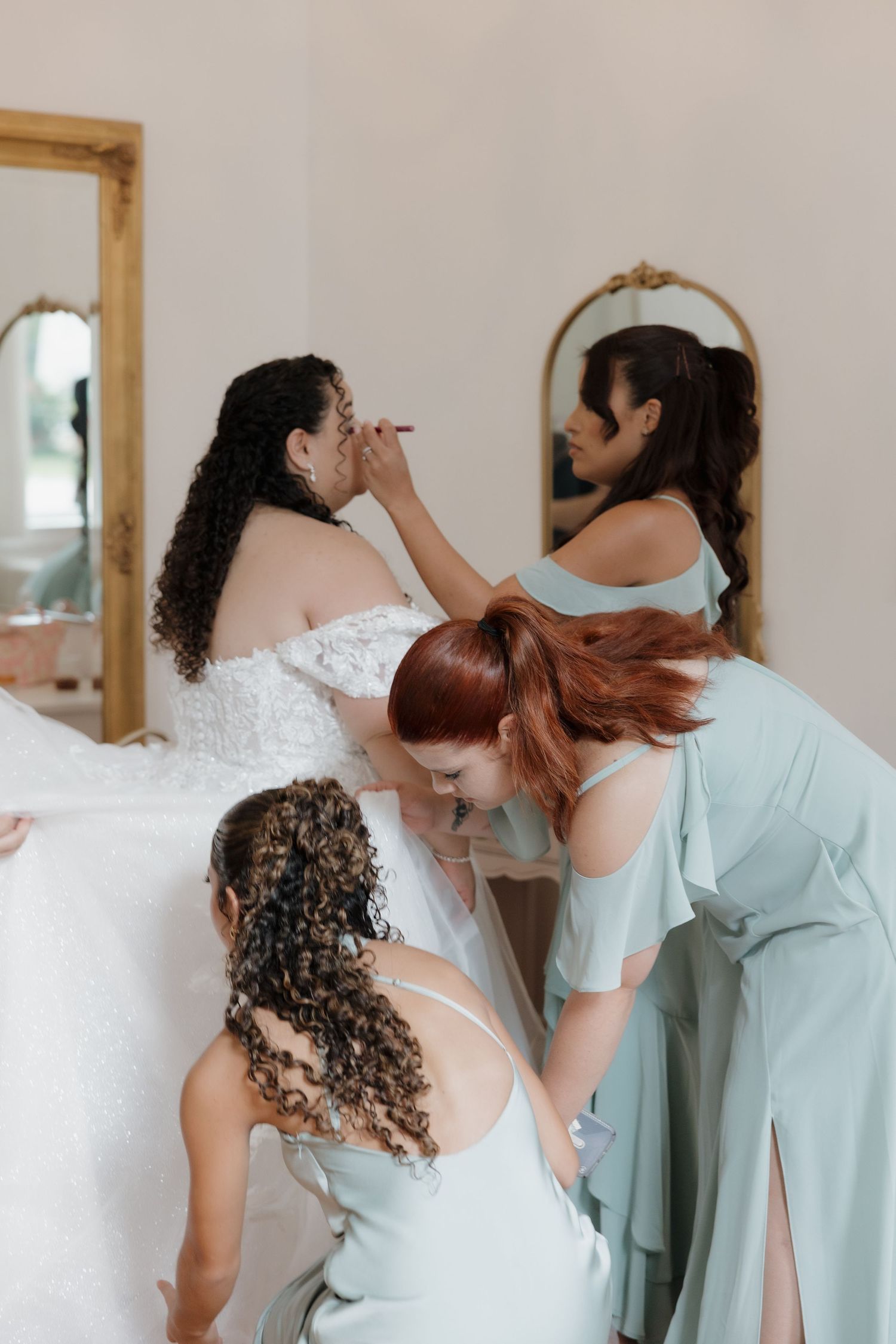 Bridesmaids in sage green dresses helping bride with makeup and dress preparations before wedding.