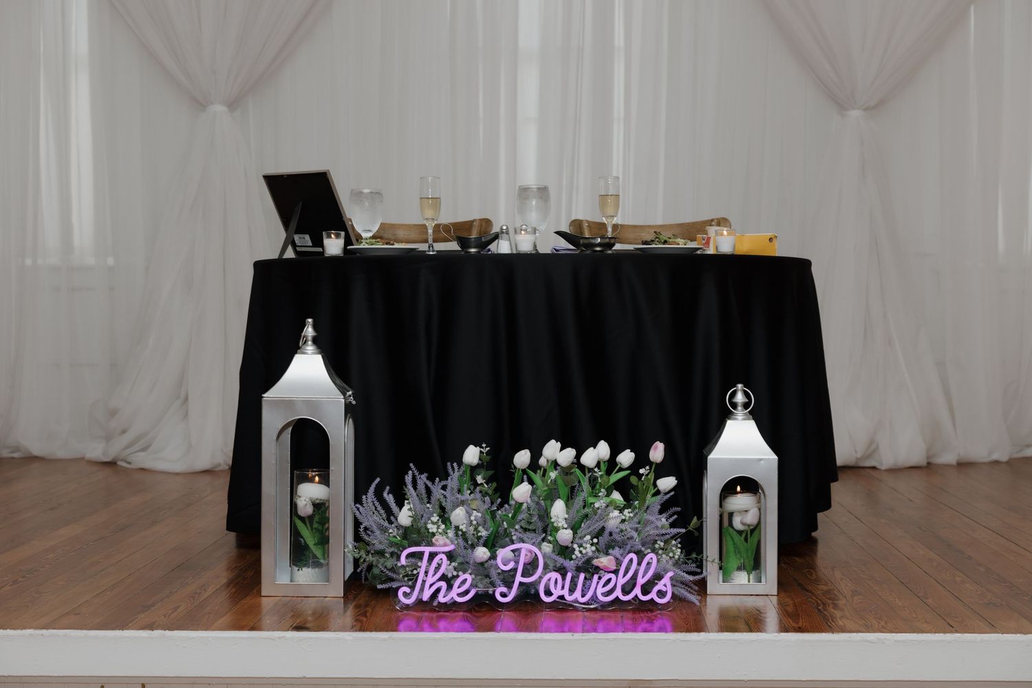 Elegant wedding sweetheart table with black linen, white lanterns, and a purple neon sign reading The Powells.