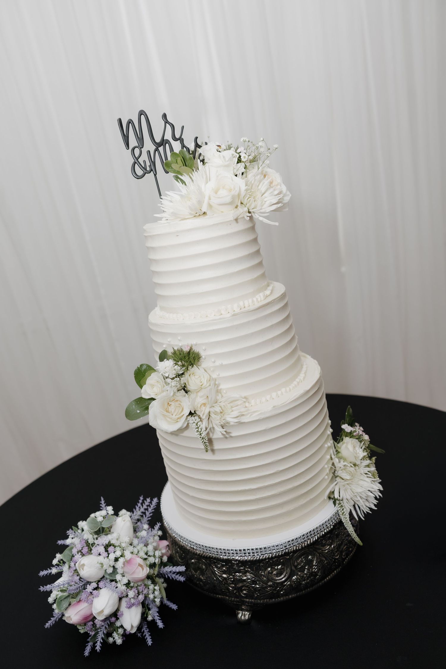 Elegant four-tier white ribbed wedding cake with floral accents and Mr. & Mrs. topper on a silver stand.