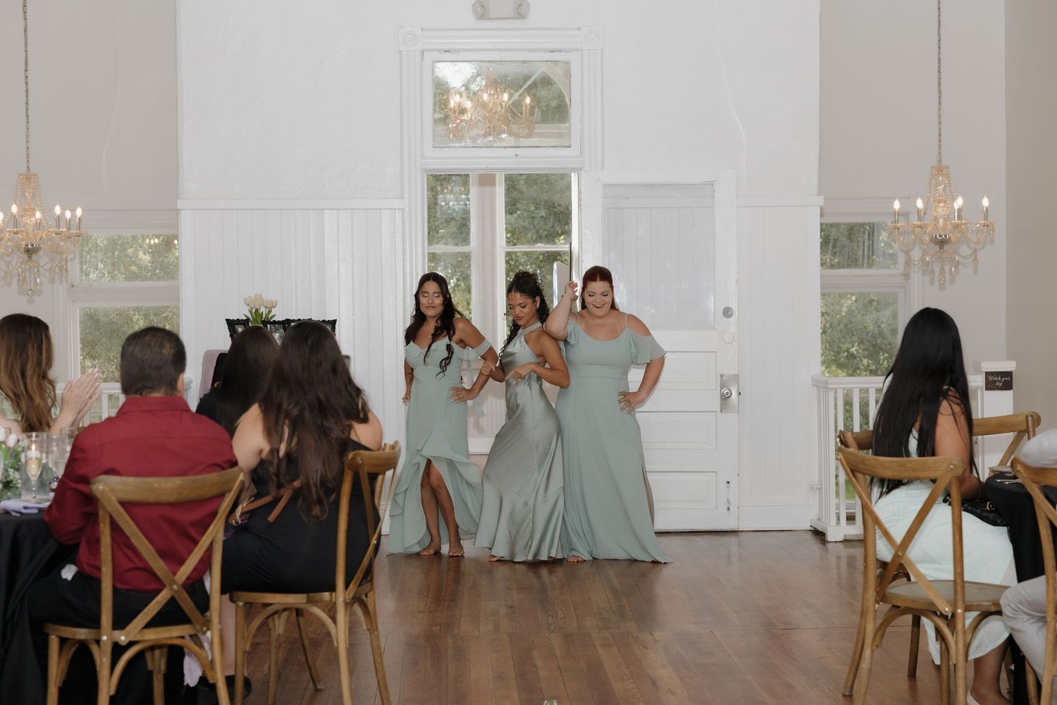 Bridesmaids in sage green dresses stand at the entrance of an elegant white wedding venue with chandeliers.