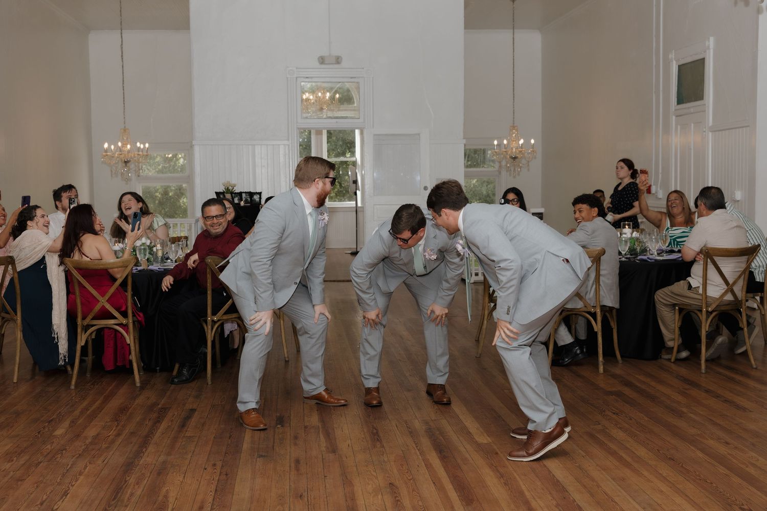 Three groomsmen in gray suits playfully bowing on a wooden dance floor at an elegant wedding reception.