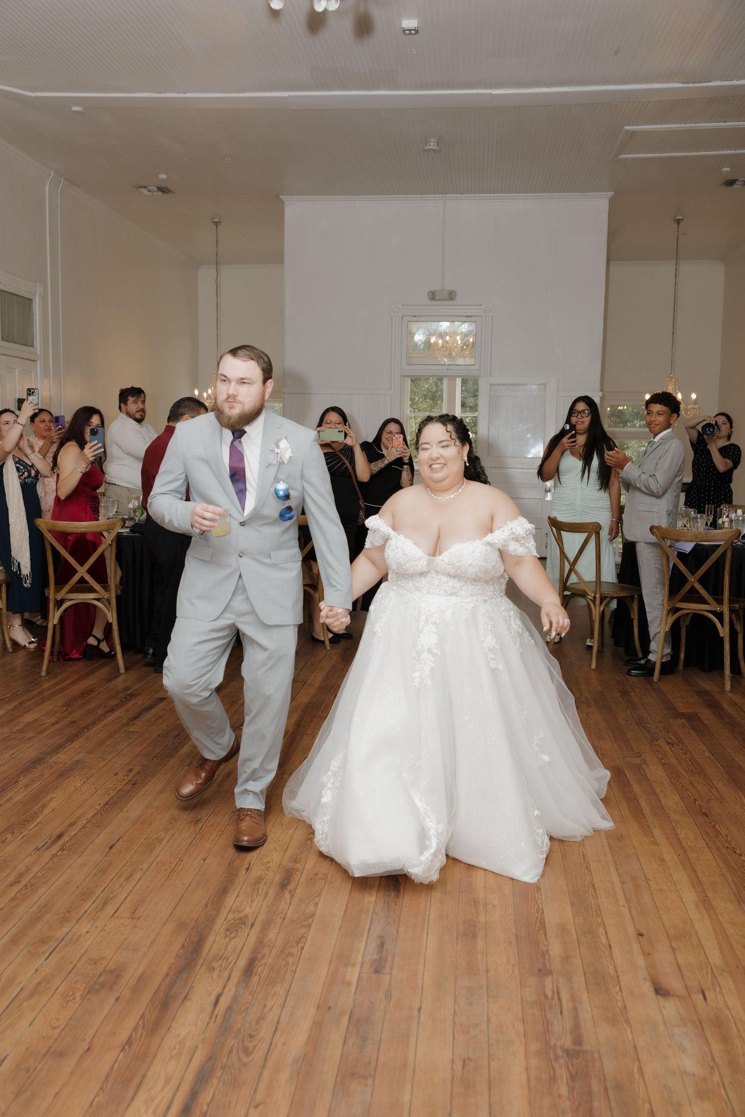 Bride and groom enter wedding reception holding hands, bride in white gown, groom in gray suit, guests celebrating.