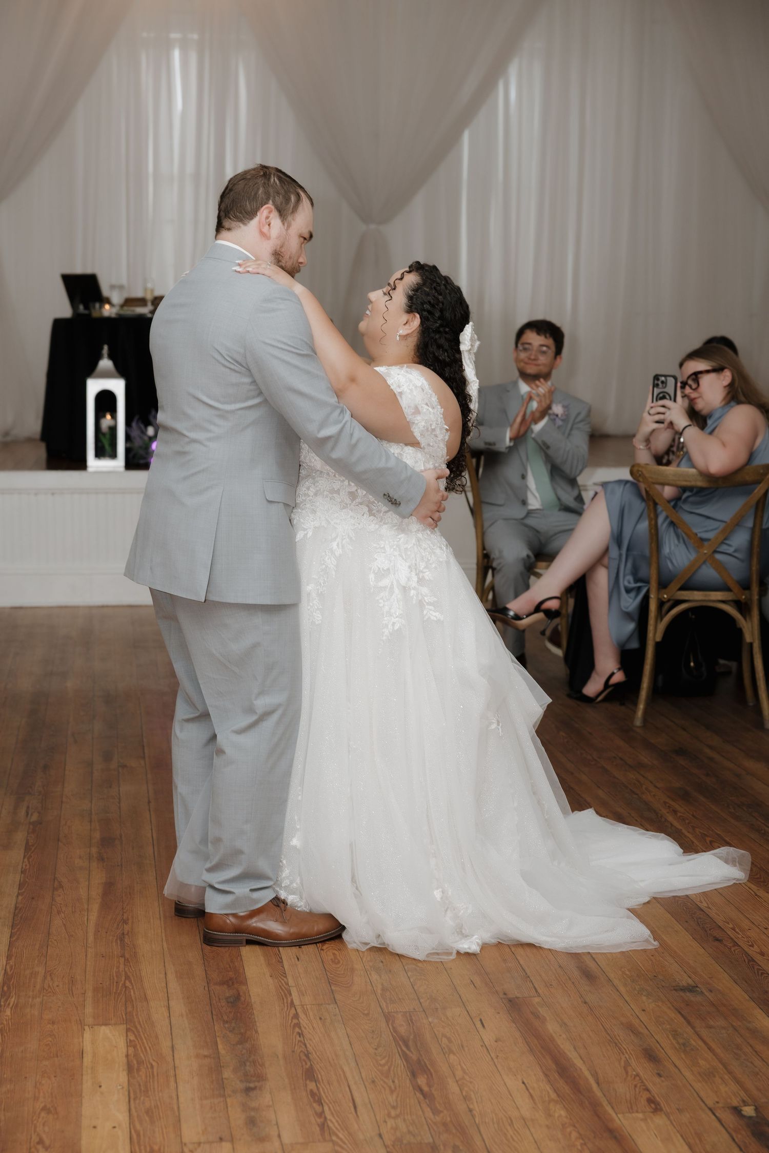 Bride and groom share their first dance at wedding reception on hardwood floor while guests watch and photograph.