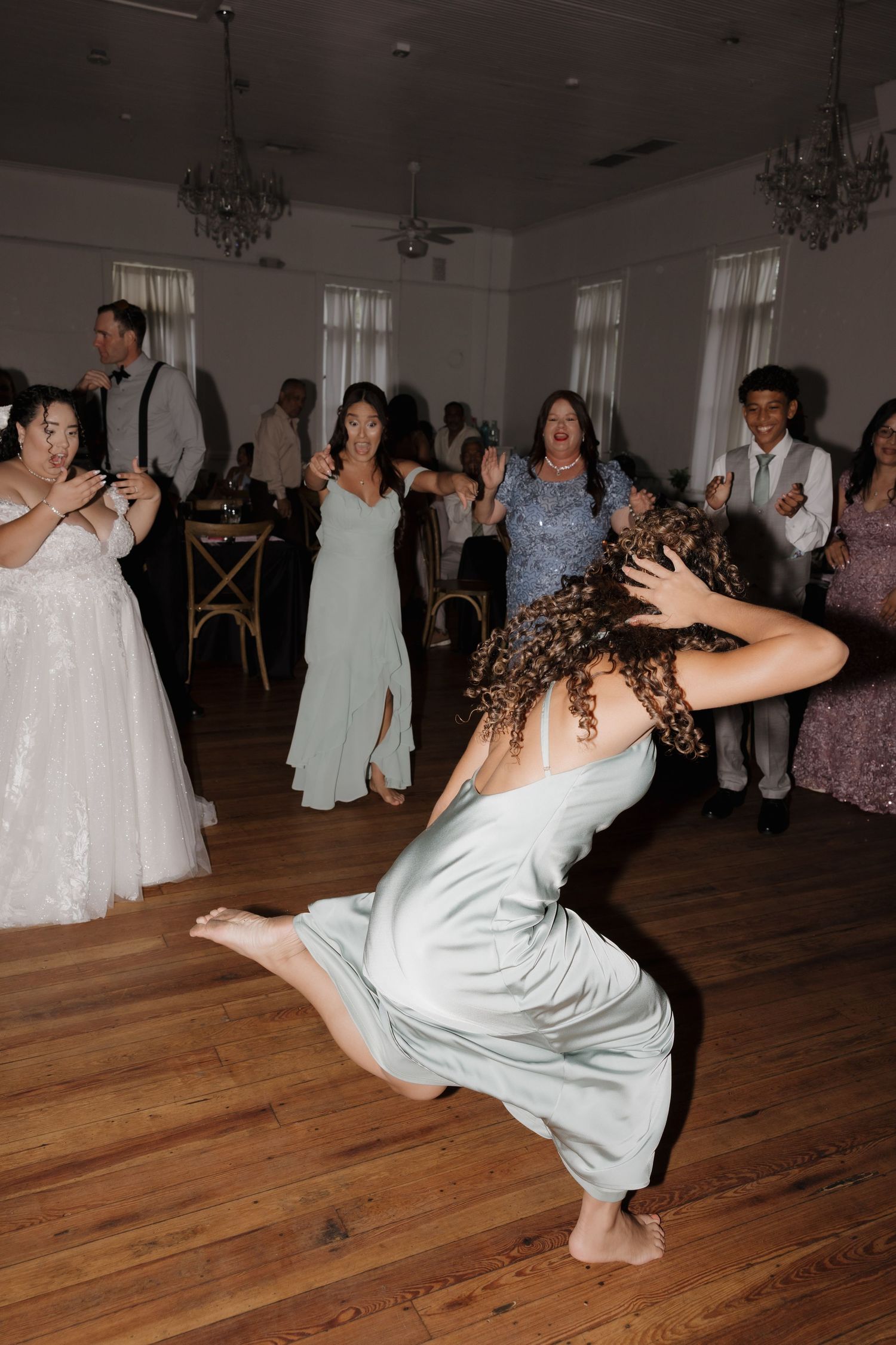 Guest in sage green dress does dramatic hair flip on wooden dance floor at wedding reception.