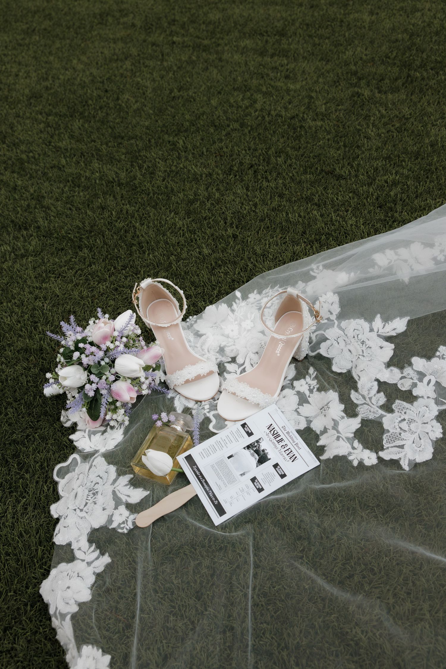 Bridal flat lay with lace veil, white heeled sandals, floral bouquet, and perfume on green grass.