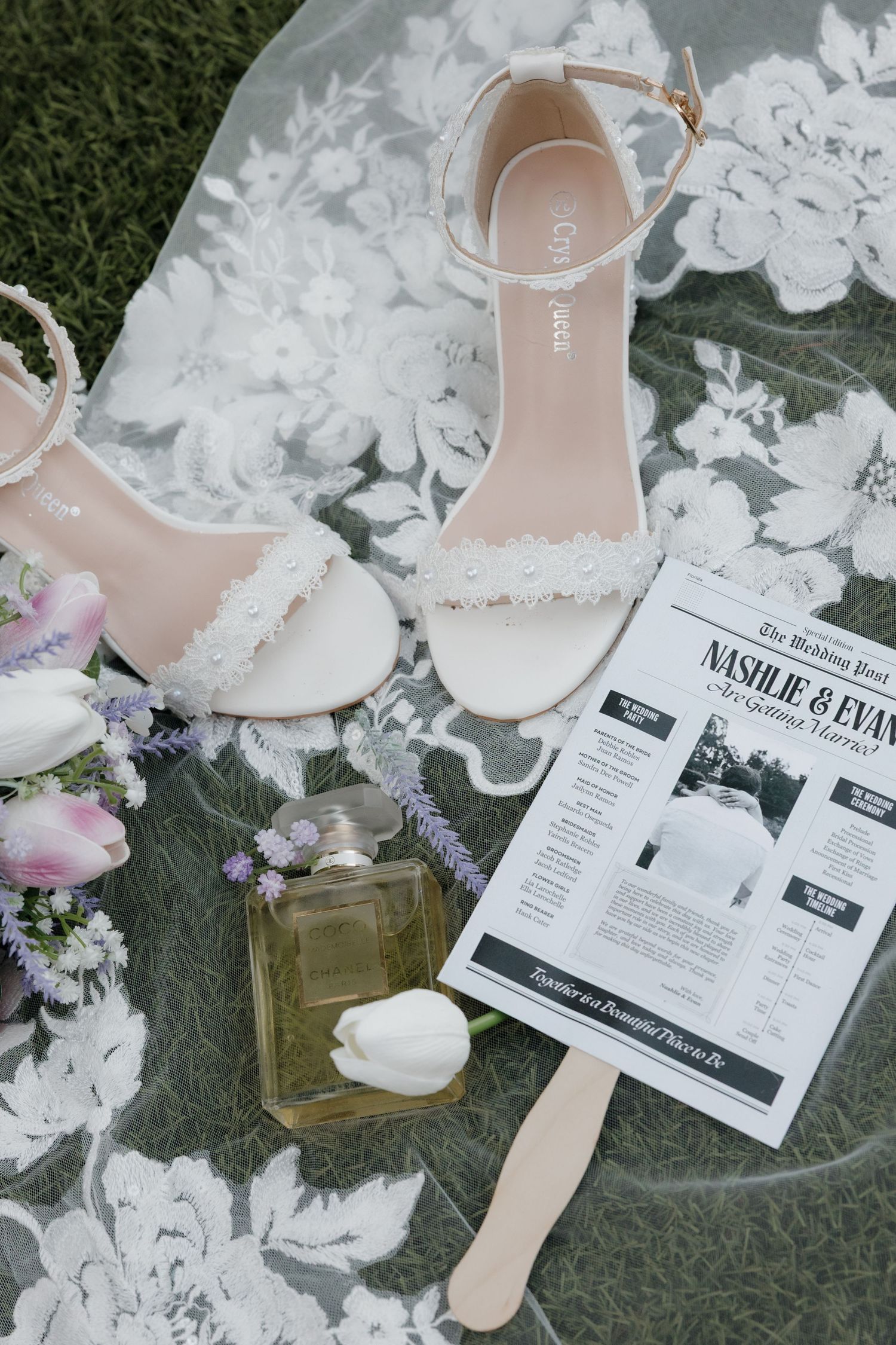 Flat lay of white lace bridal heels, perfume, tulips and wedding program newspaper on floral fabric.