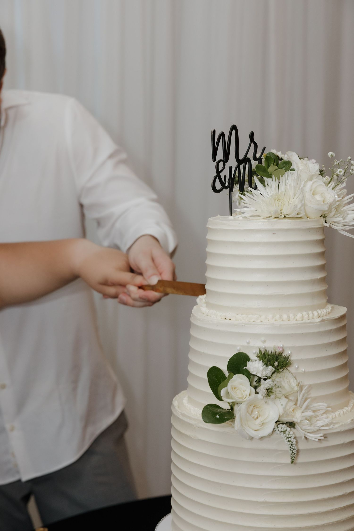 Three-tier white wedding cake with Mr & Mrs topper, white floral decorations, being cut by person in white shirt.