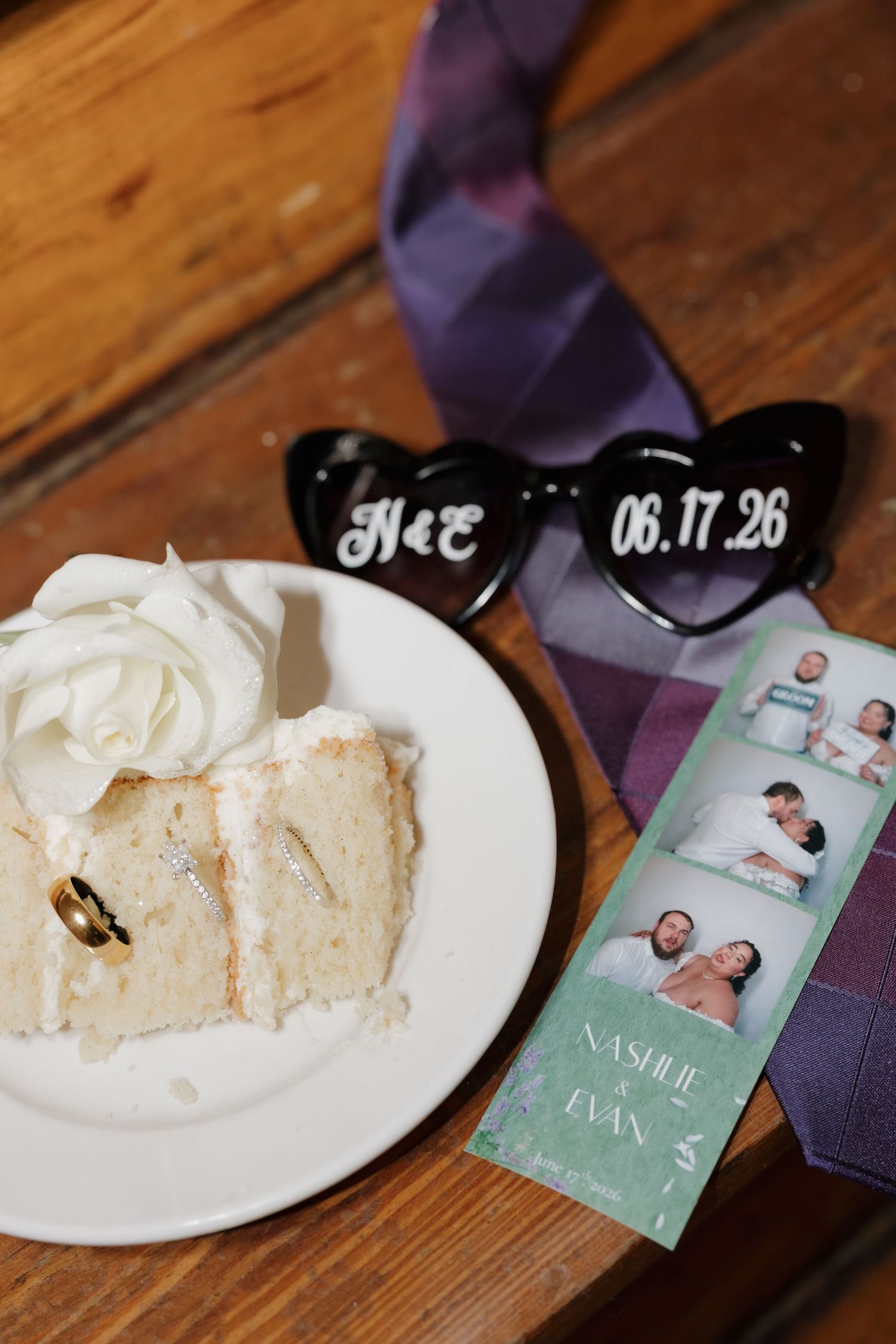 Wedding flat lay with cake slice, rings, heart sunglasses, photo strip, and purple tie on wood.