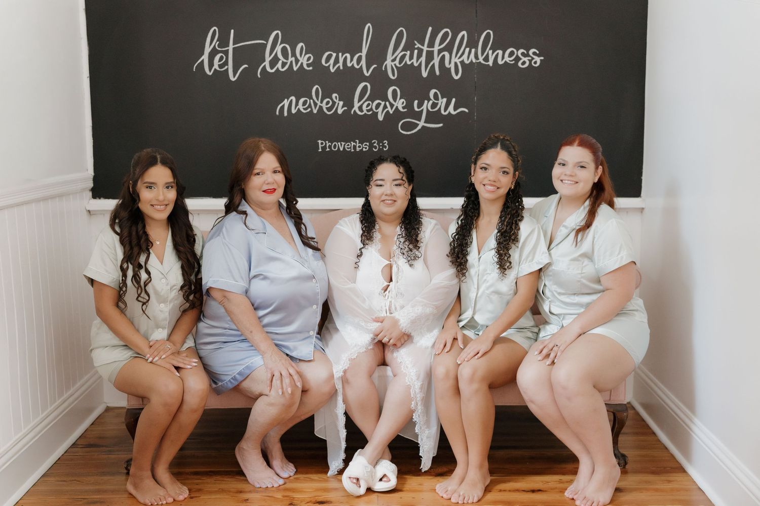 Five women in matching robes sitting on bench beneath faith quote chalkboard wall sign.