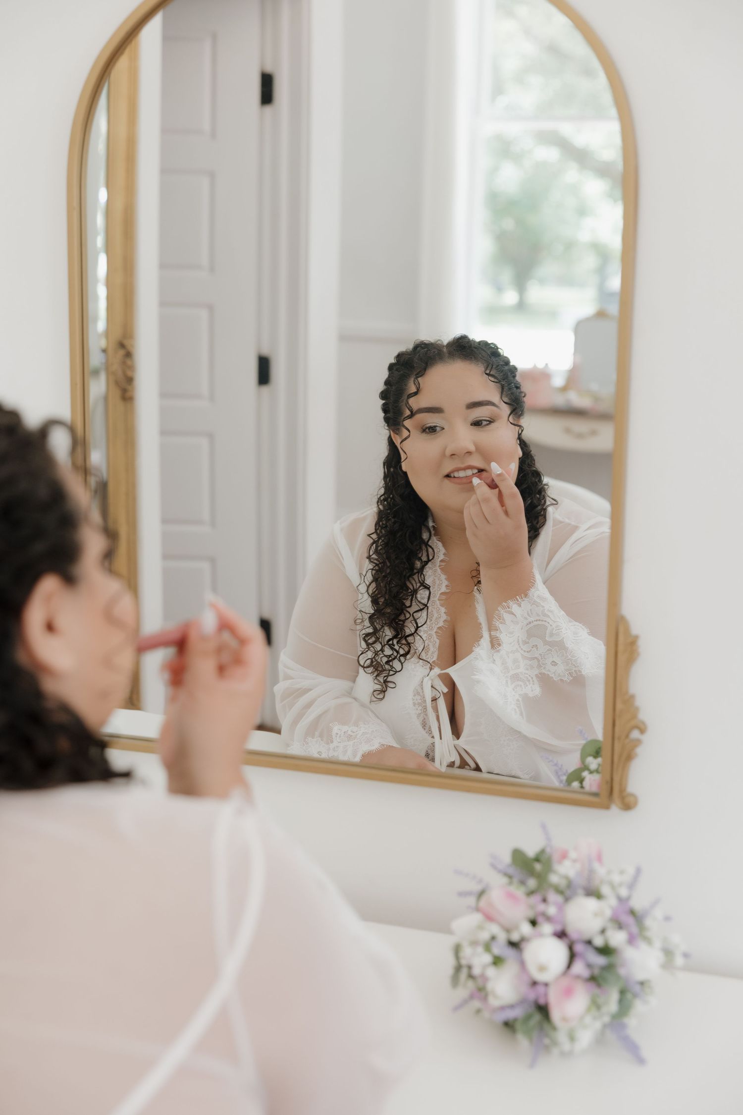 Bride in white lace robe applies lipstick while gazing into gold arched mirror on wedding morning.