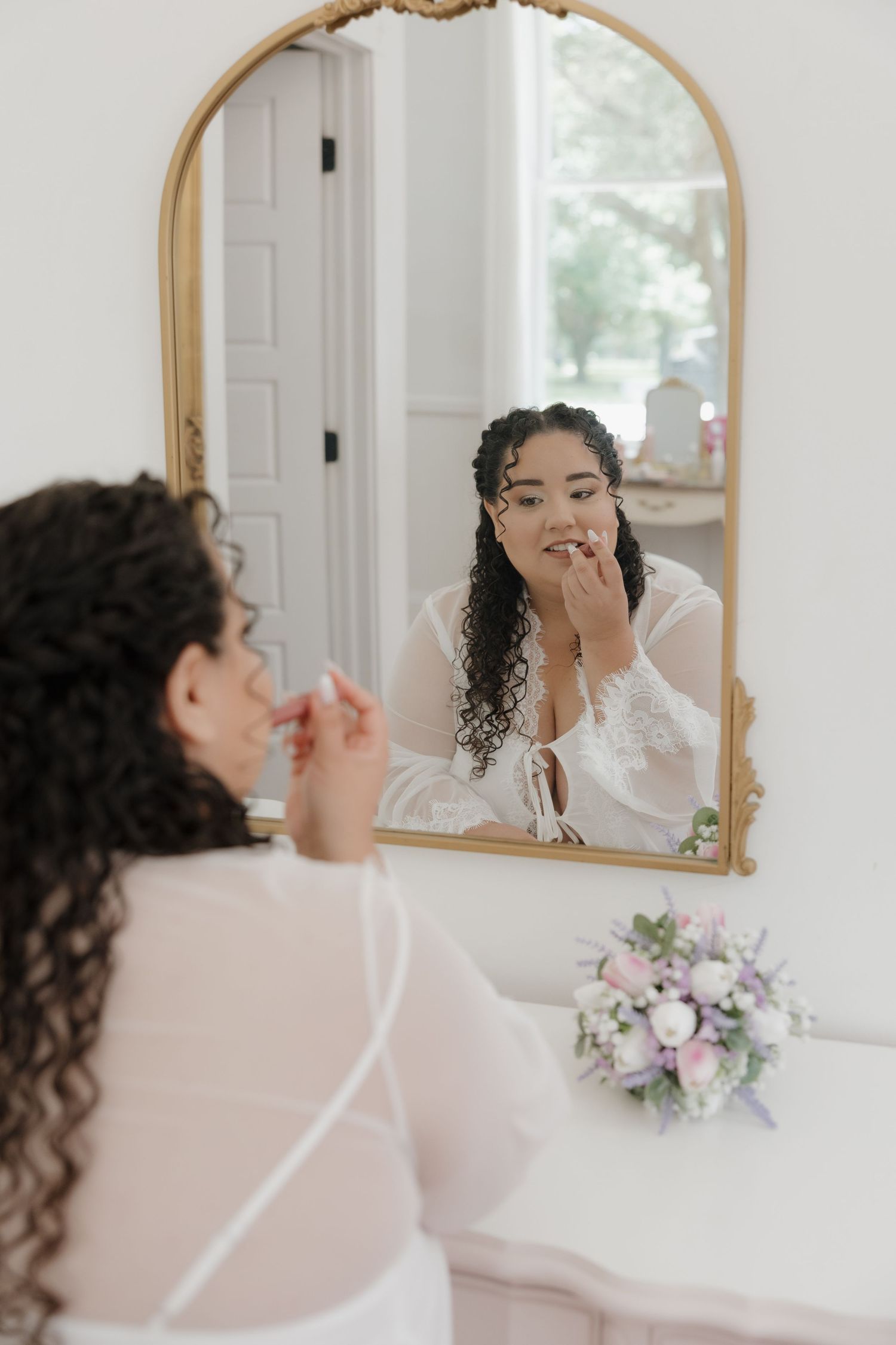 Bride with curly hair applies lipstick while looking into ornate gold arched mirror on wedding day.