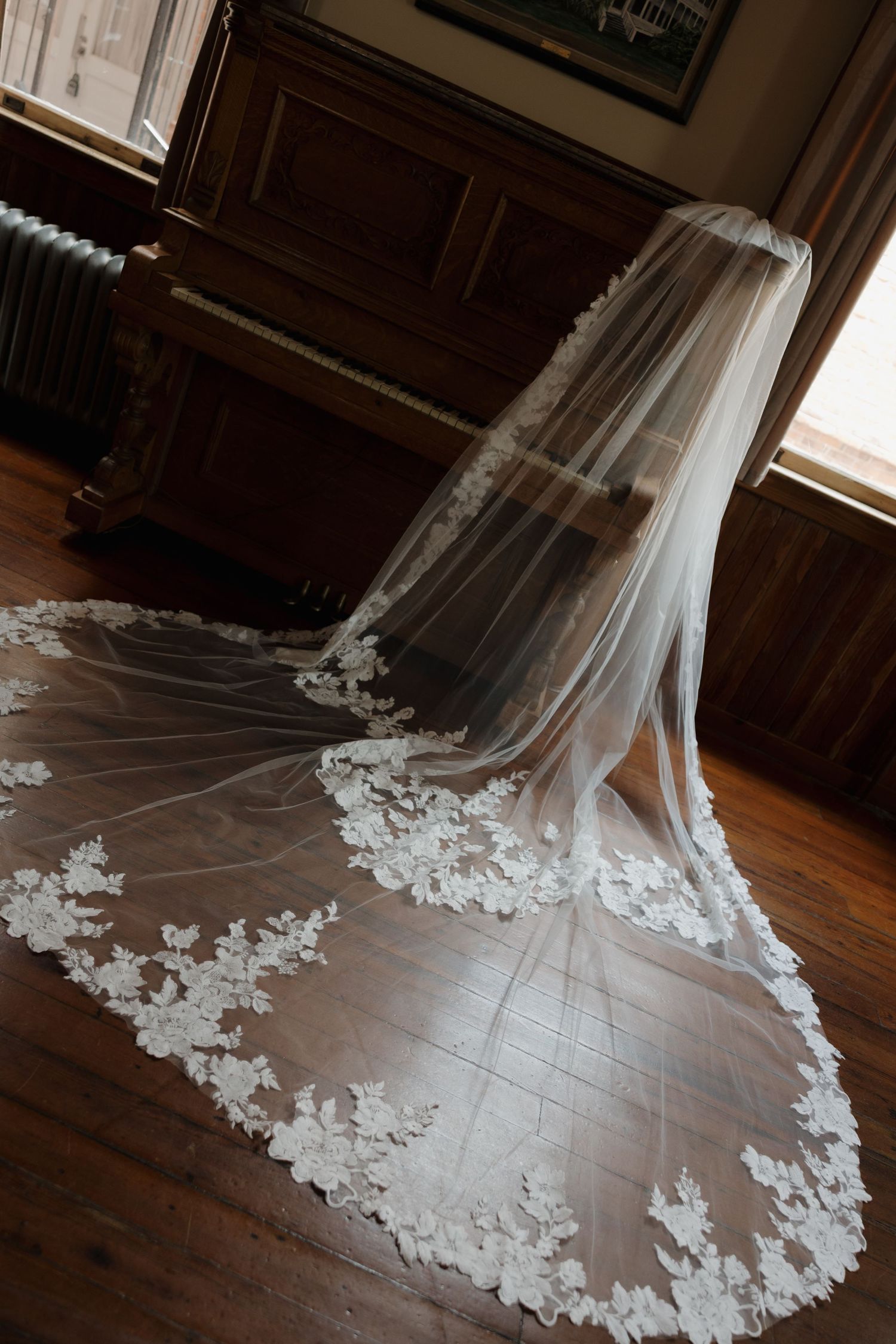 Elegant lace-trimmed cathedral wedding veil draped on hardwood floor near vintage piano in dimly lit room.