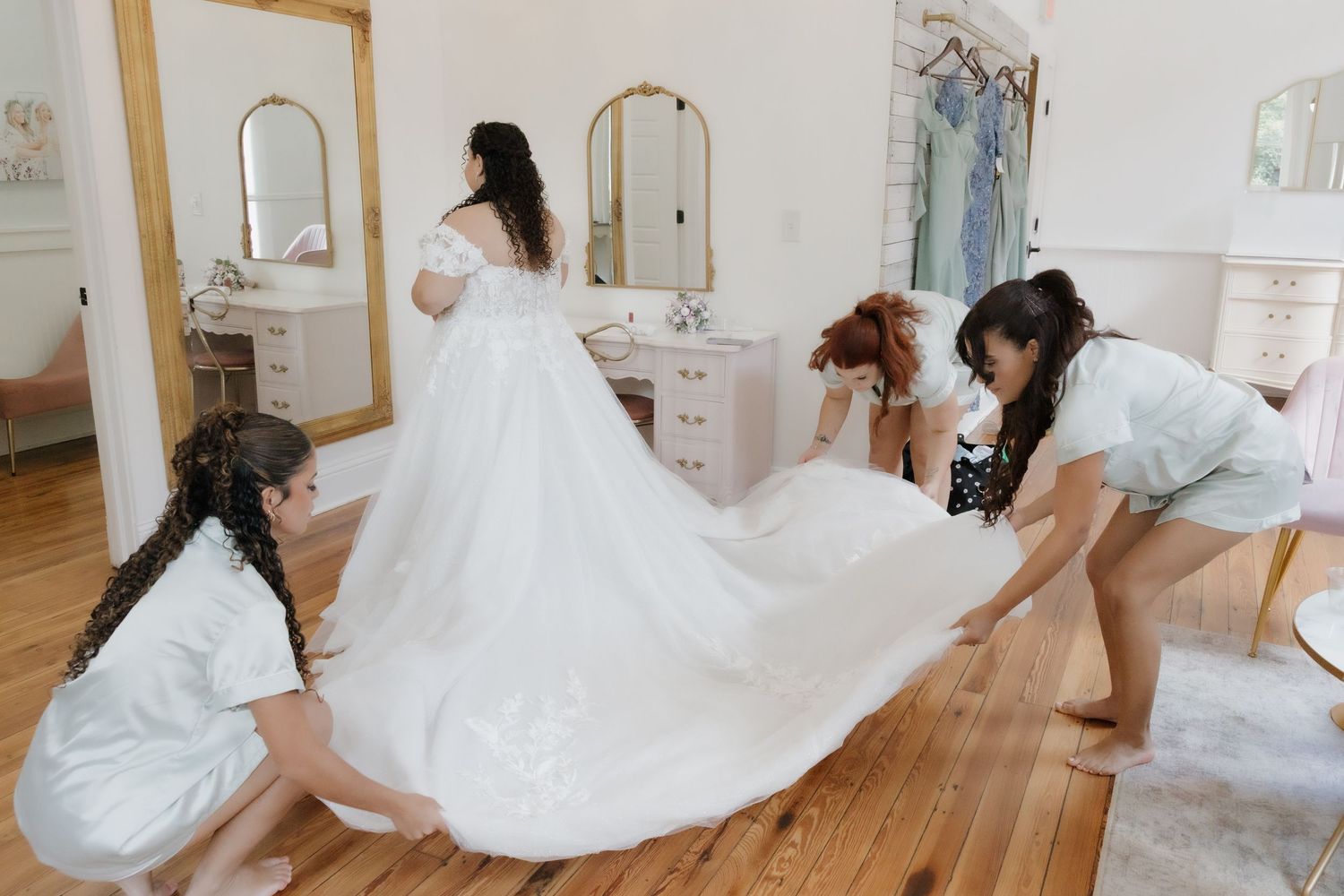 Bridesmaids in sage green dresses help arrange bride's flowing white wedding gown train in bridal suite.