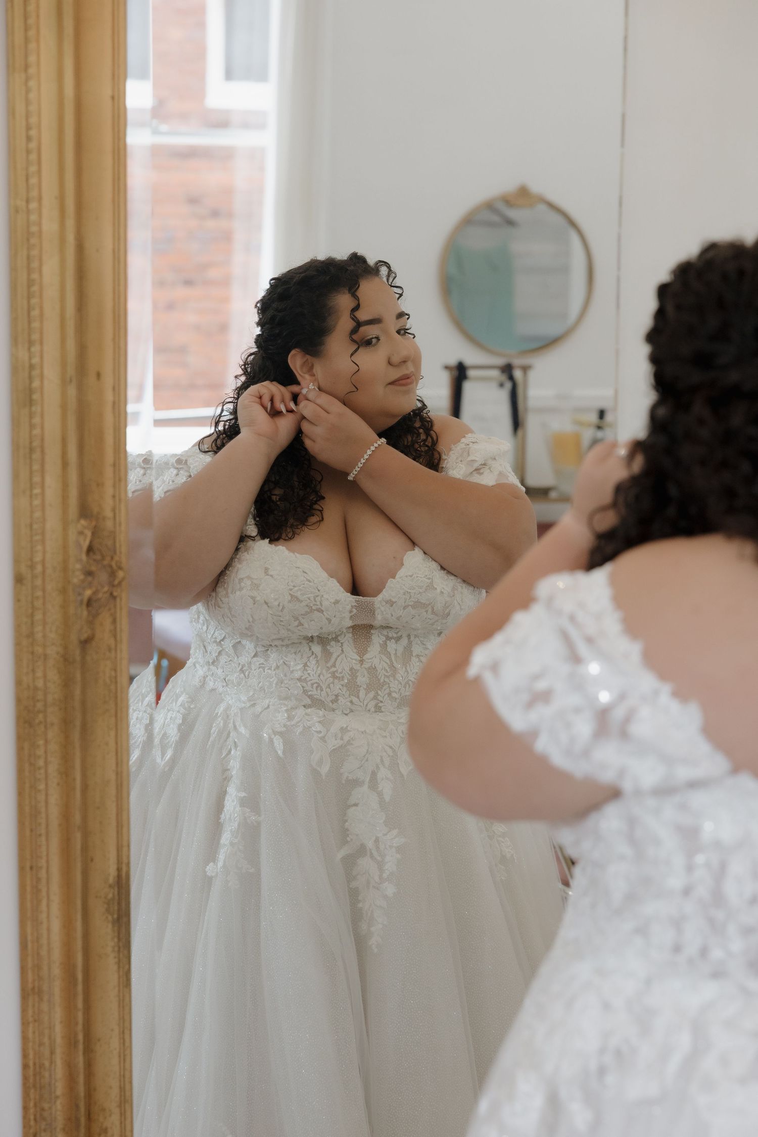 Plus size bride in off-shoulder lace wedding gown adjusting earrings in ornate gold mirror.