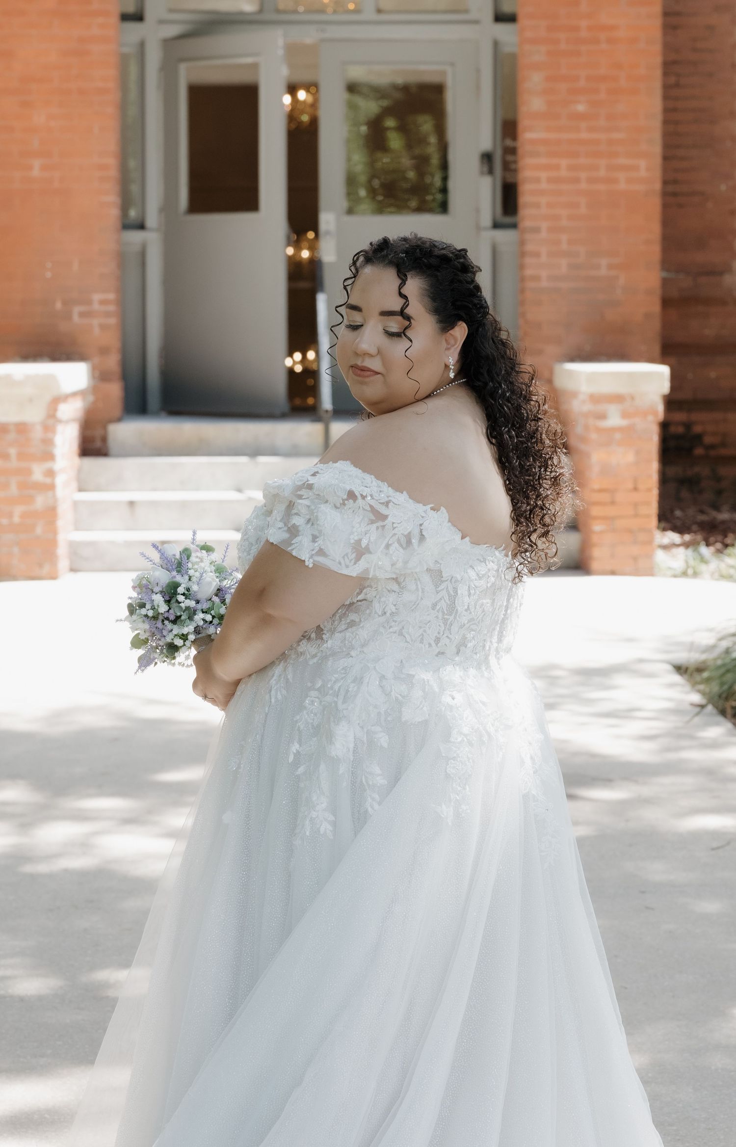 A bride in an off-shoulder lace wedding gown holds a bouquet outside a brick building.
