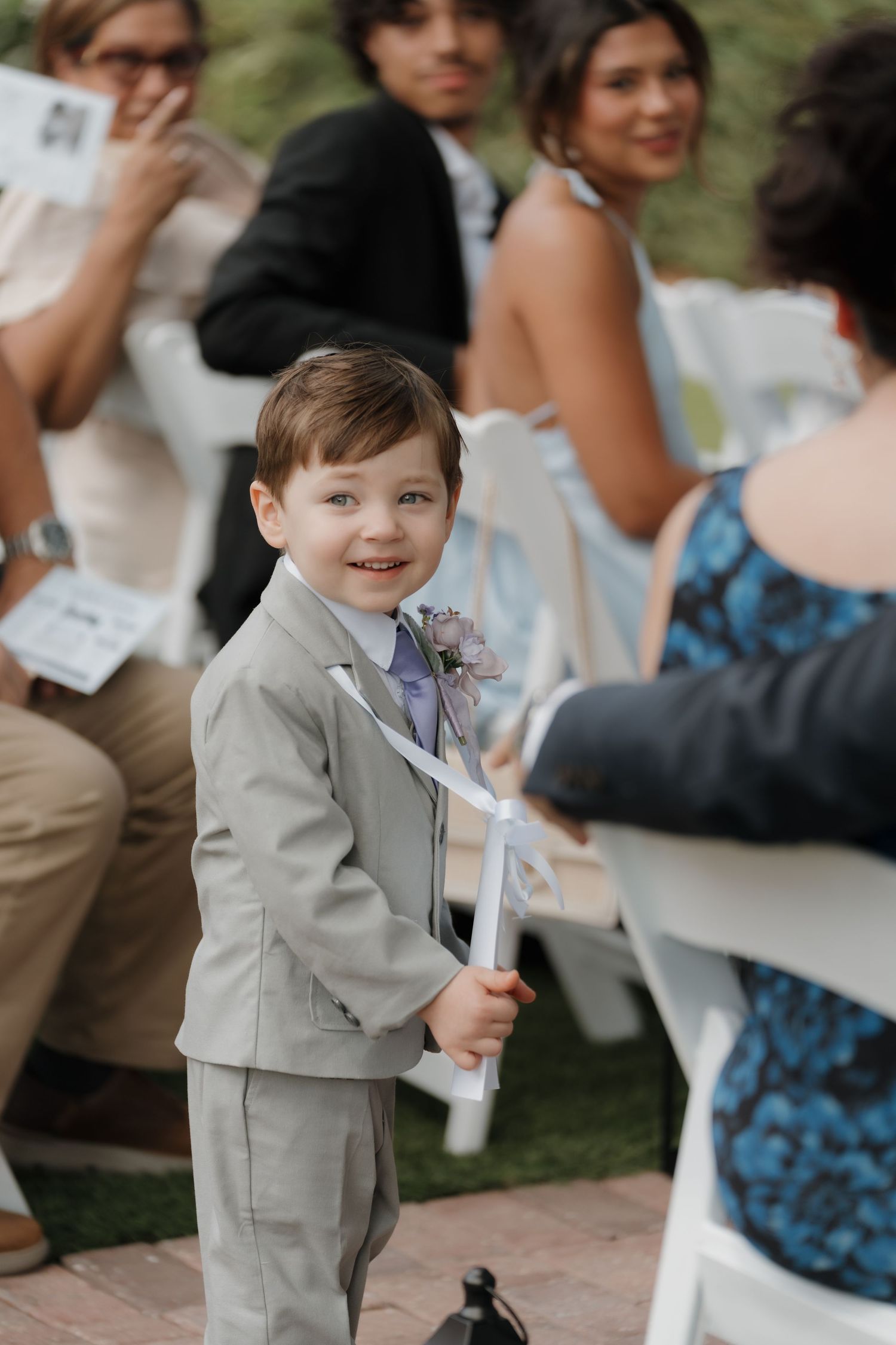 Adorable young ring bearer in gray suit smiles at camera during outdoor wedding ceremony.