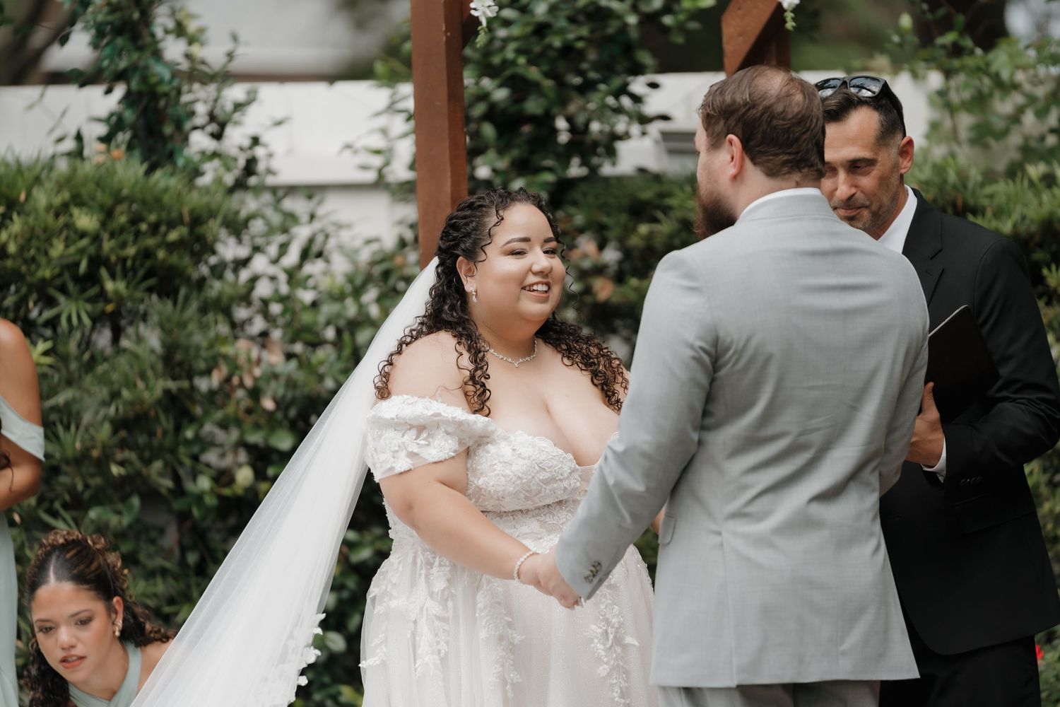 Joyful bride in off-shoulder lace gown smiling at groom during outdoor wedding ceremony.