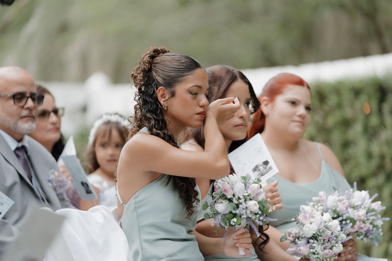Emotional bridesmaid wiping tears during outdoor wedding ceremony holding bouquet of flowers.