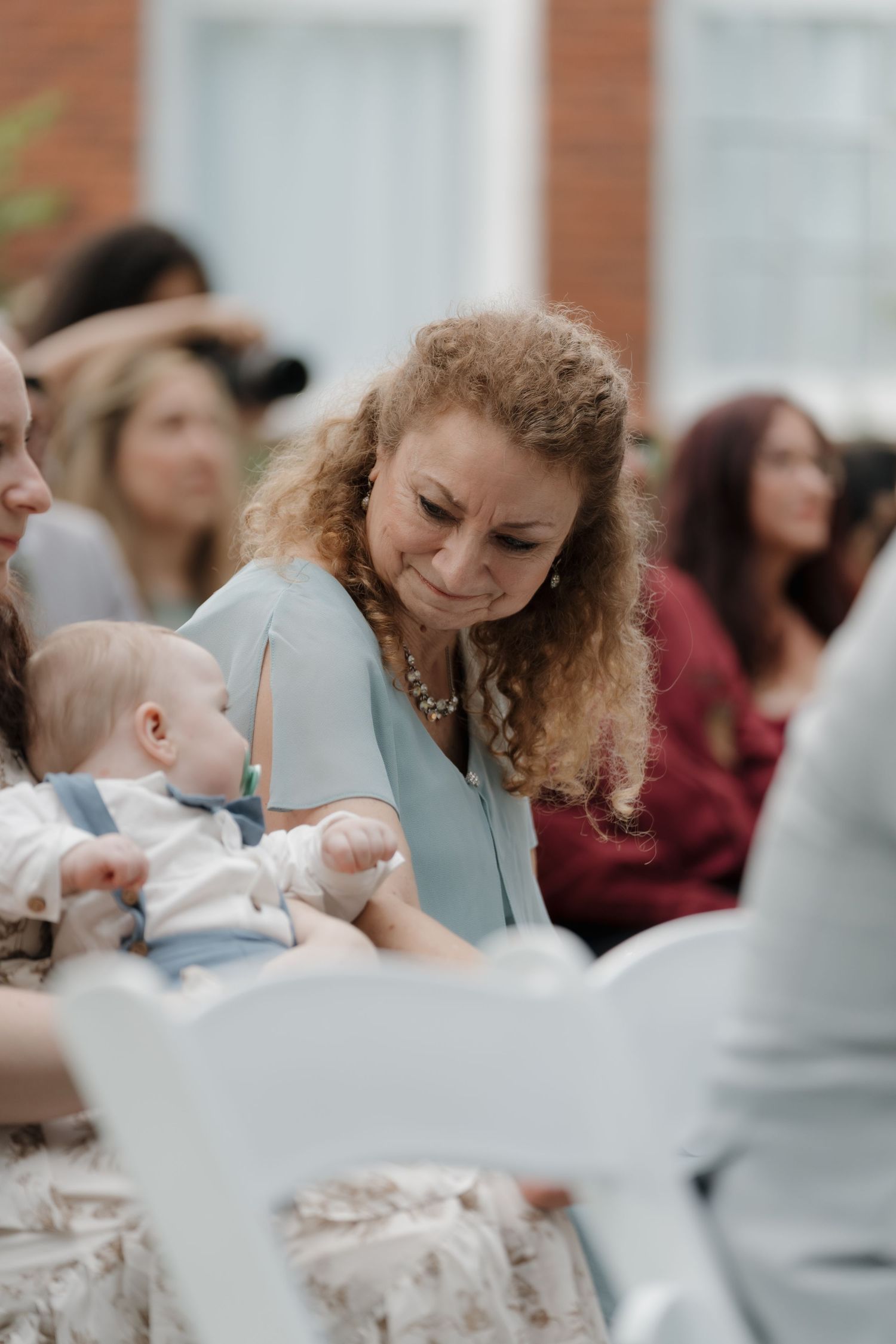 A woman with curly hair in a light blue dress smiles at a baby during an outdoor wedding ceremony.