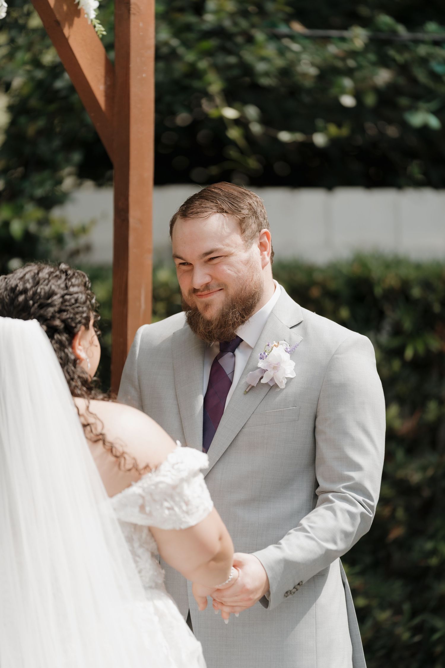 Bearded groom in grey suit with purple tie smiling at bride during outdoor wedding ceremony.