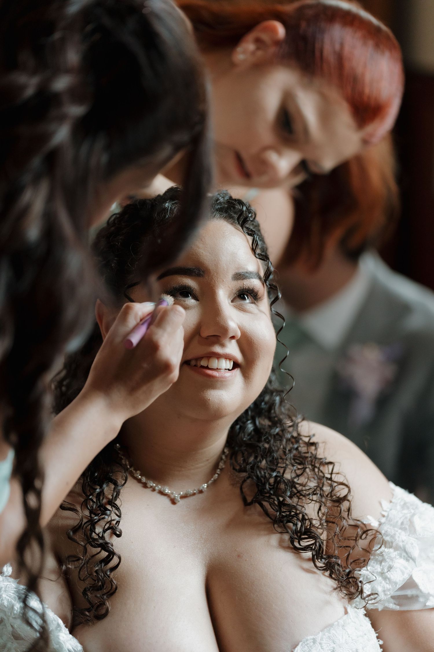 A smiling bride with curly hair gets makeup applied by a redheaded bridesmaid in a white lace wedding dress.