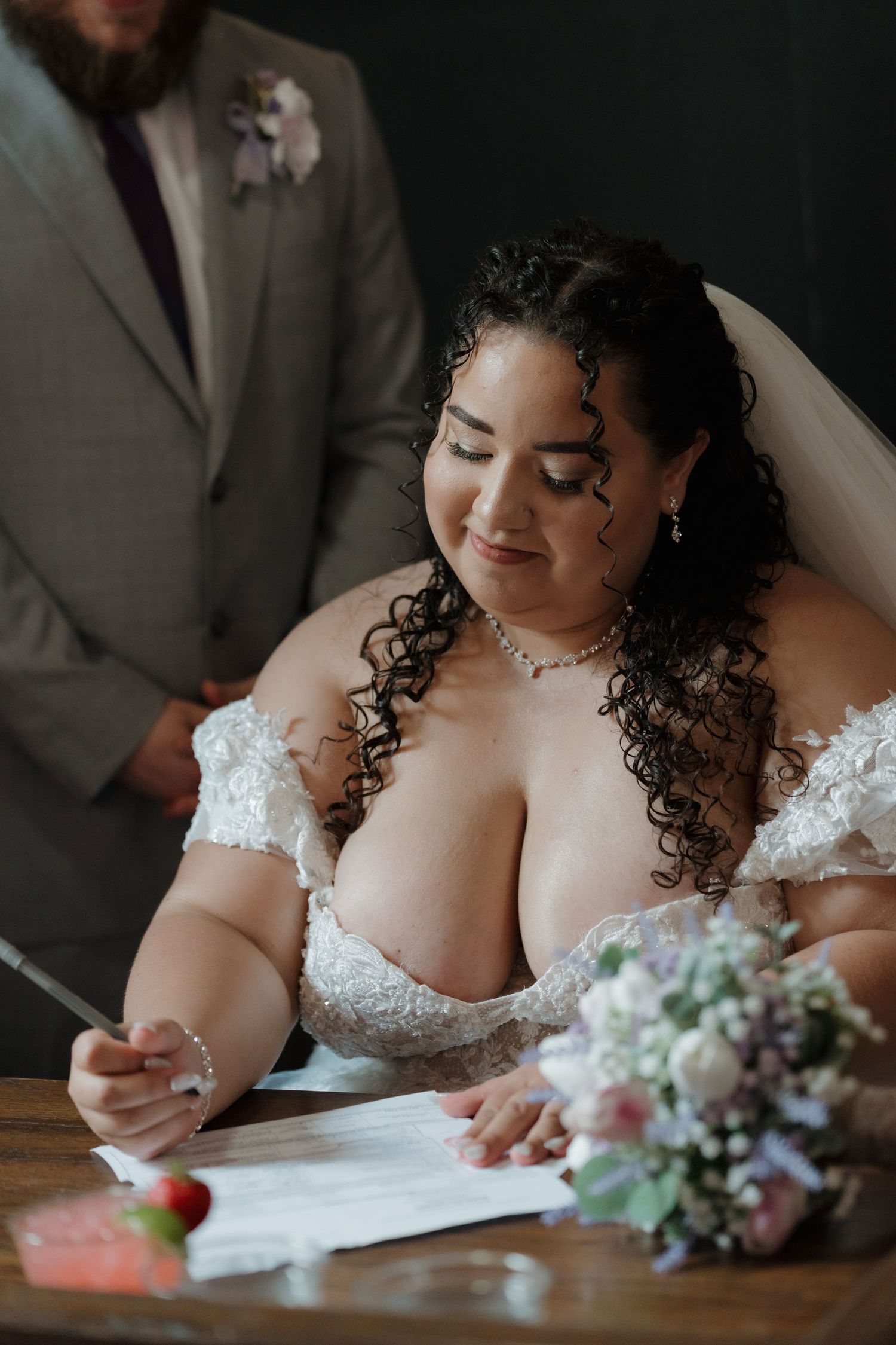 Bride in lace off-shoulder wedding gown signing marriage certificate at wooden table with floral bouquet nearby.