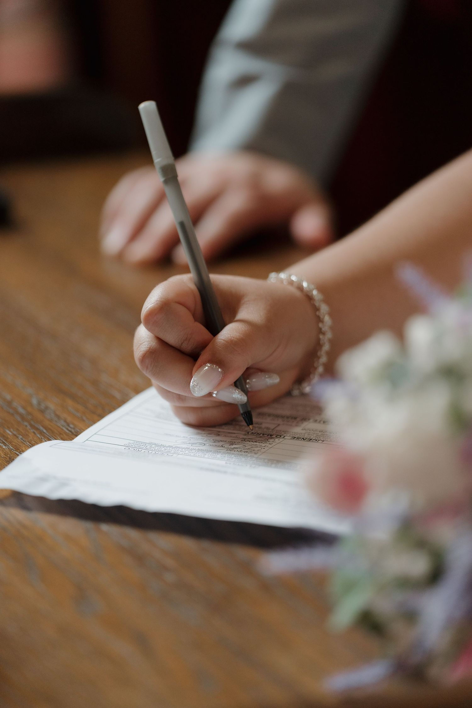 Bride signing wedding certificate with pearl bracelet and French manicure, pen in hand at wooden table.