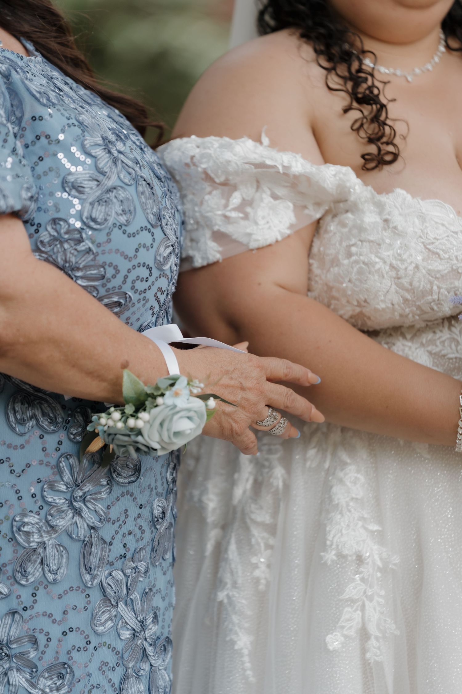Bride in white lace gown stands arm-in-arm with mother wearing blue sequin dress and floral wrist corsage.