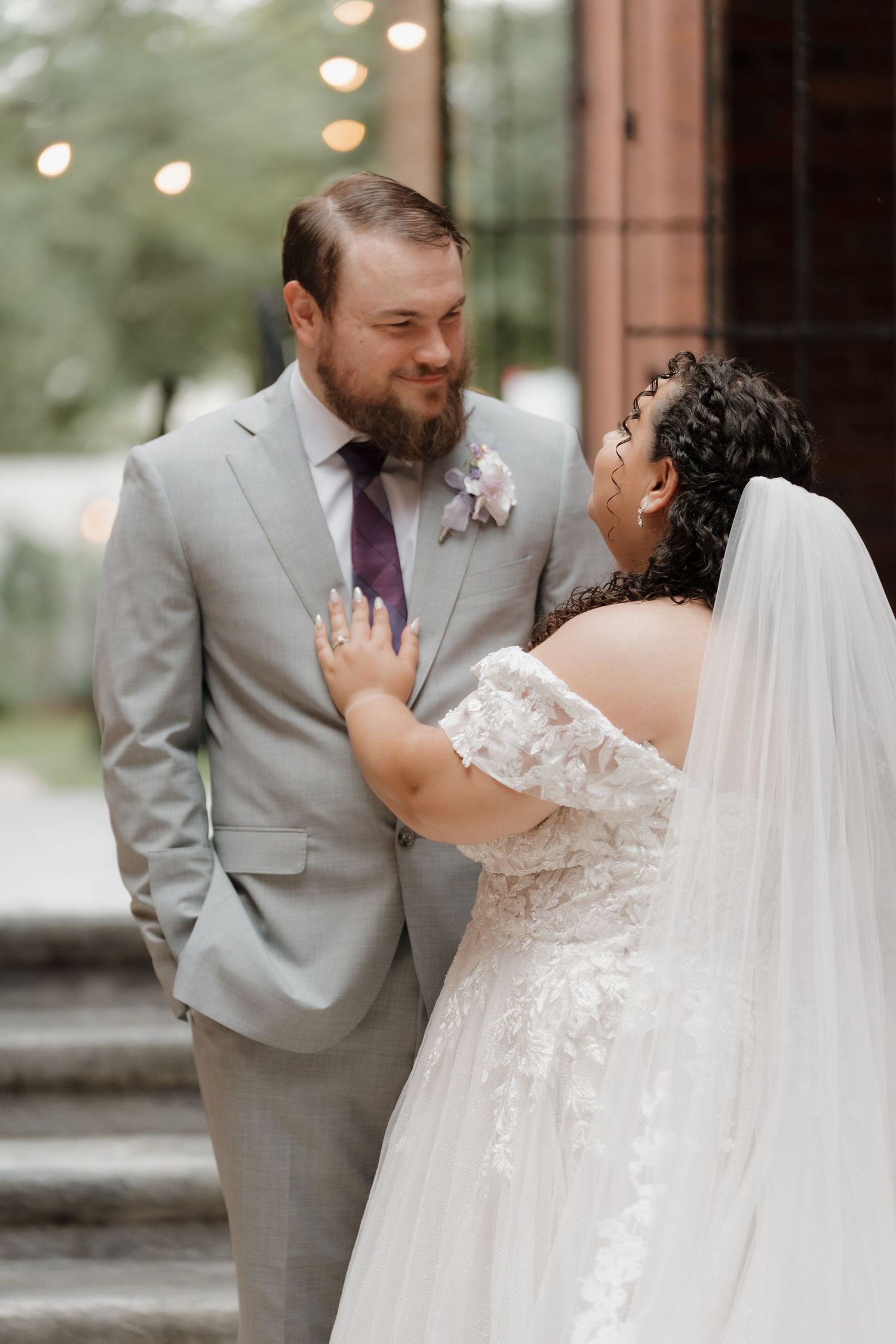 Bride in off-shoulder lace gown laughs joyfully with bearded groom in gray suit and purple tie.