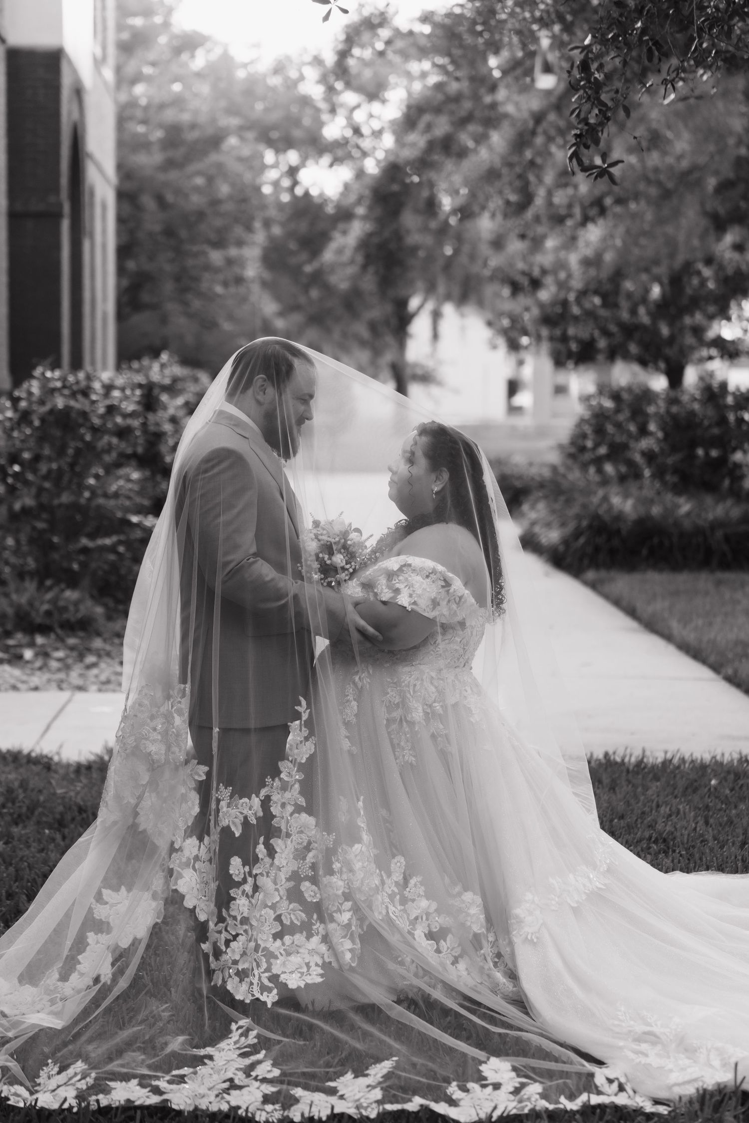 Black and white photo of bride and groom sharing romantic moment under flowing lace wedding veil outdoors.