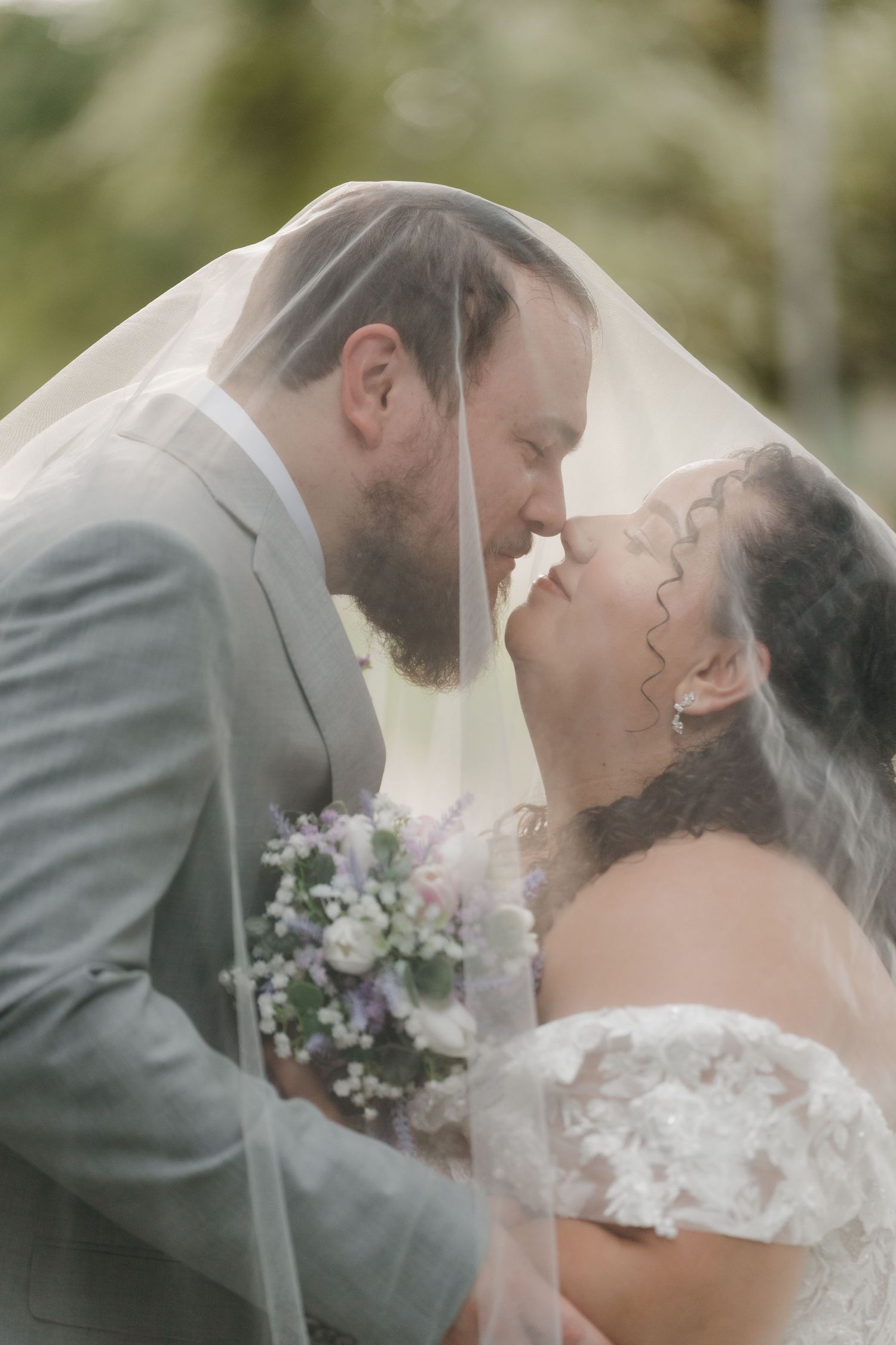 Bride and groom share a tender kiss under sheer wedding veil while holding purple and white bridal bouquet.