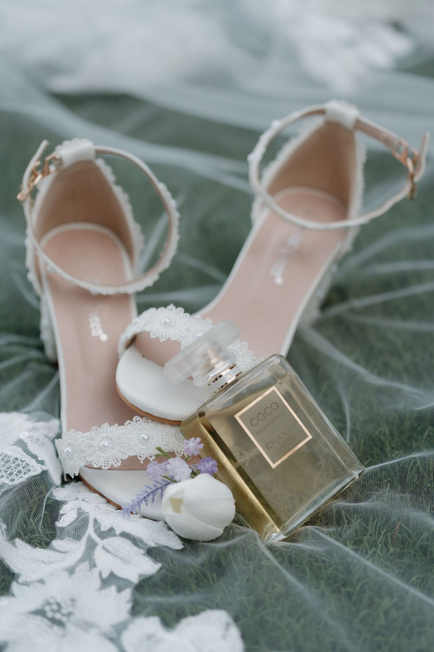 White bridal heels with lace details beside a Chanel perfume bottle and white tulip on sage green fabric.